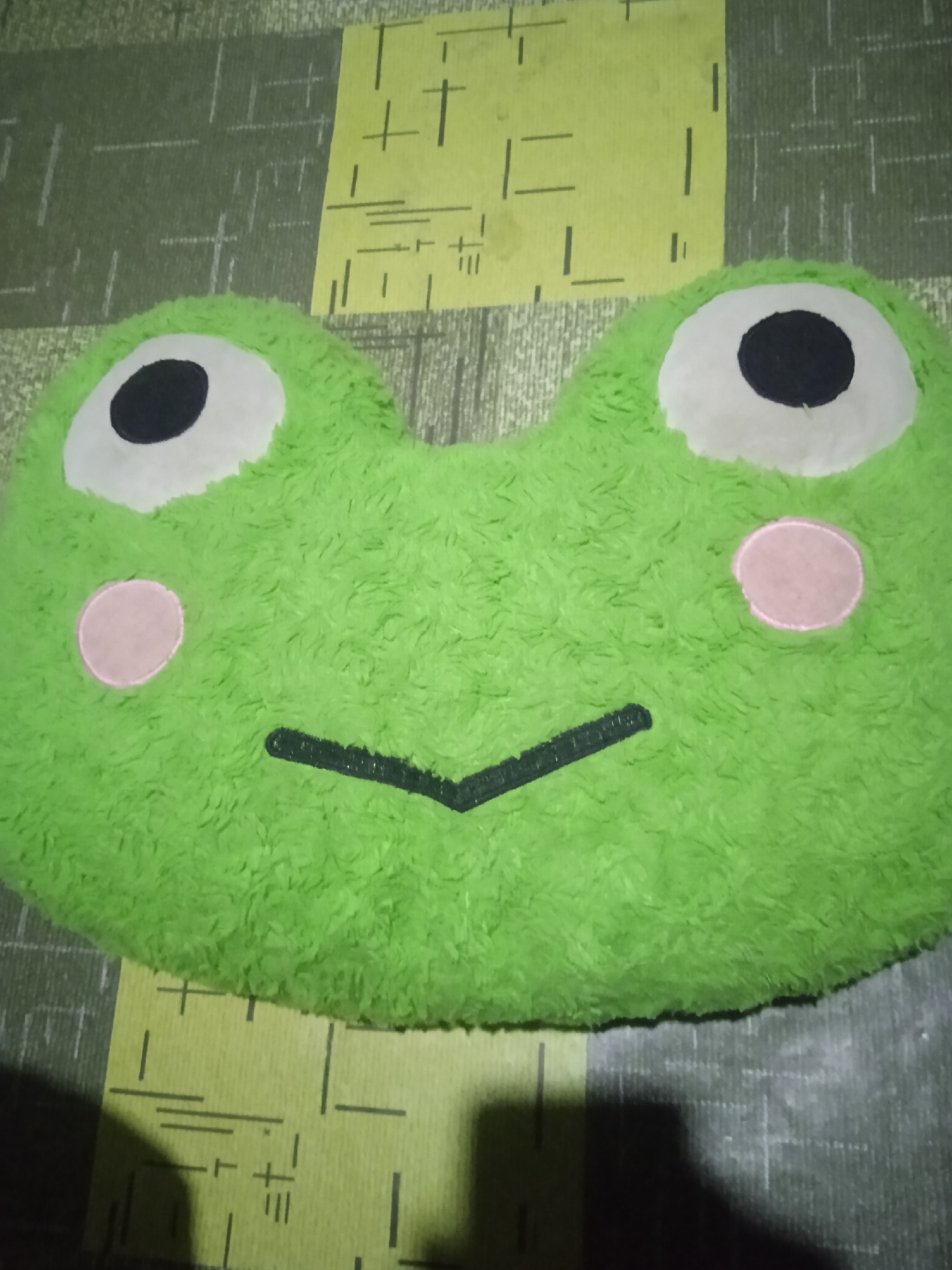 A green plush pillow shaped like a frog's head rests on a surface with a patterned fabric. The pillow has large white eyes with black pupils, and two pink circles on its cheeks. A black line forms its mouth. The patterned surface beneath the pillow consists of alternating green and yellow rectangular sections with thin white lines and some black markings. The image is taken from a slightly overhead angle, with the pillow dominating the frame. The lighting appears to be dim, casting some shadows. The specific location within Bora, Indonesia, is not ascertainable from the visual information provided.