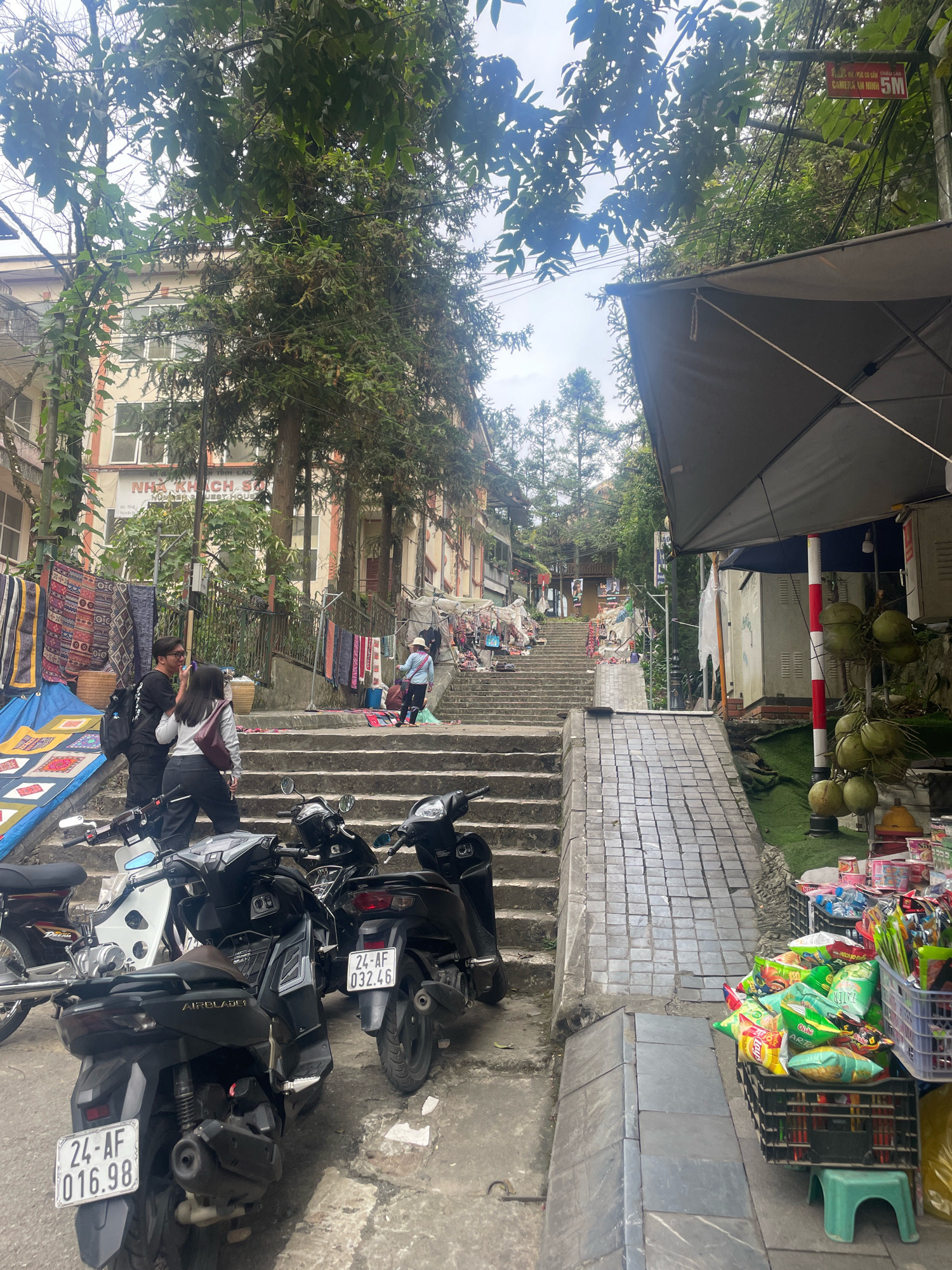The image depicts a narrow street scene in Sa Pa Ward, Vietnam, characterized by a steep set of stone stairs leading upwards. The stairs are flanked by buildings on the left and a vendor stall on the right. Several motorcycles are parked on the lower part of the street. A couple, a man and a woman, are standing on the stairs, facing each other and appearing to interact. The woman is wearing a grey sweater and black pants, with a burgundy bag slung over her shoulder. The man is wearing a black t-shirt and black pants, carrying a backpack. Further up the stairs, a person in a blue shirt and hat is walking. The vendor stall on the right displays a variety of goods, including packaged snacks and coconuts, with a large umbrella providing shade. The architecture suggests a blend of traditional and modern elements, with foliage and trees integrated into the urban environment. A sign reading "NHÀ KHÁCH SO" and "NUMBER GUEST HOUSE" is visible on a building. The overall impression is of a busy pedestrian and commercial area.