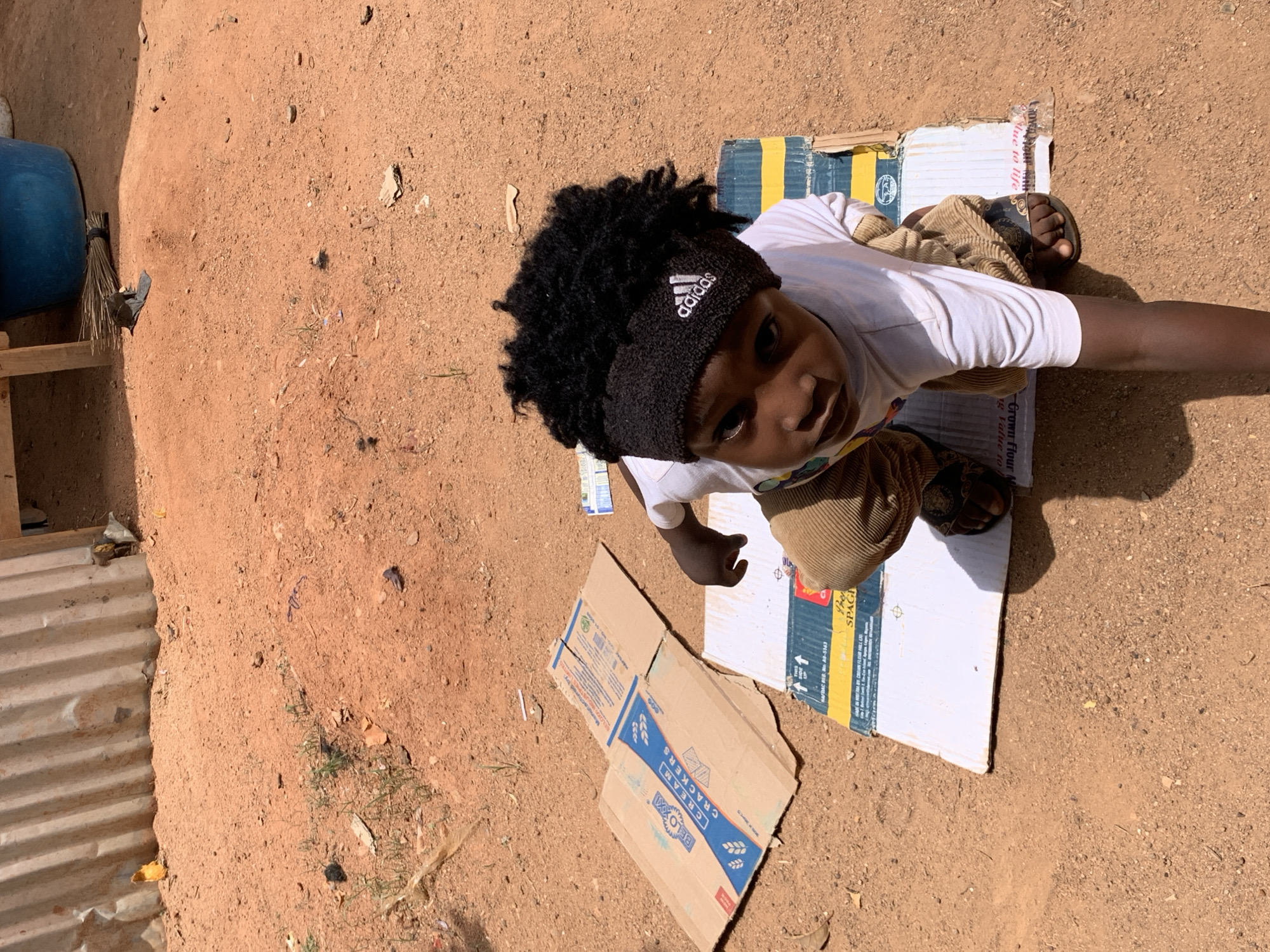 A young individual is lying on several pieces of cardboard on a dirt surface. They are wearing a white shirt, brown corduroy pants, and black sandals. The person also has a black Adidas headband. A blue plastic container, a wooden structure, and corrugated metal are visible in the background. Parts of a cardboard box for "Beloxxi Cream Crackers" are visible on the ground. The background appears to be a dirt yard or open space. It is assumed that this image was taken in Rafin Bauna, Nigeria.