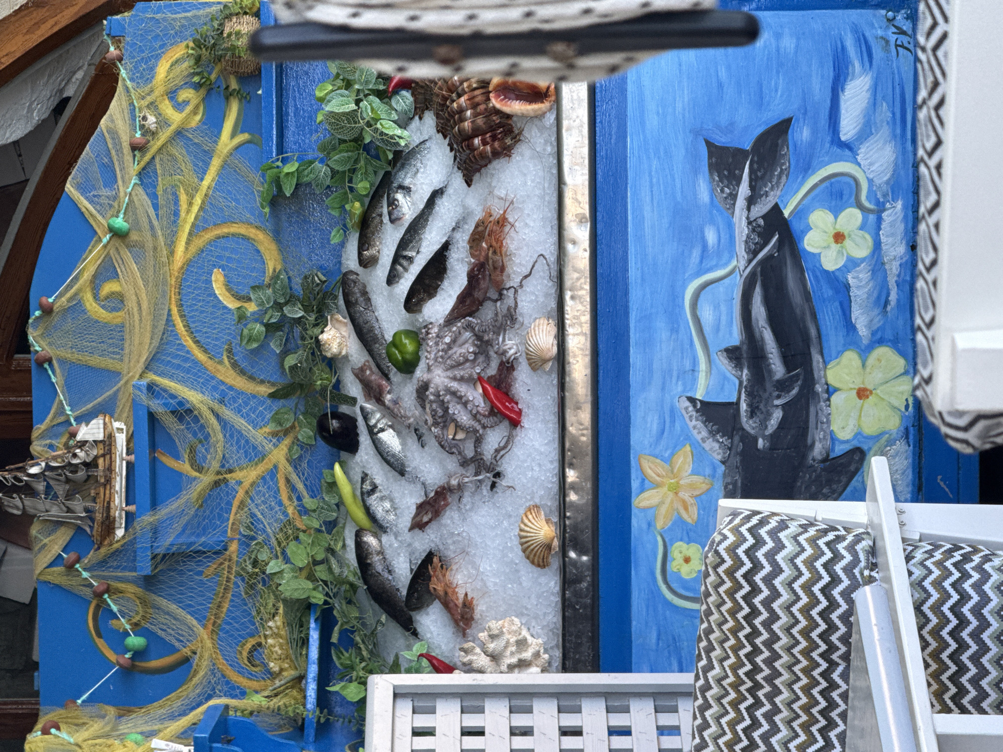 The image depicts a display of fresh seafood, likely in a restaurant or market, in Rethymno, Greece. The display is set against a vibrant blue background adorned with nautical-themed decorations, including a model ship, fishing nets, and faux greenery. The seafood, arranged on a bed of ice, includes various fish, shrimp, and an octopus. Some of the fish appear to be whole and unscaled, while others are filleted.
In the lower part of the image, a mural on the blue background features a stylized shark swimming amongst flowers and clouds. The overall scene suggests a seaside establishment that emphasizes fresh catches. The presence of outdoor seating with patterned cushions indicates it's an open-air or semi-outdoor setting. There are no people visible in the frame. The image's composition and subject matter strongly suggest a location in a coastal area known for its seafood, such as Rethymno.
