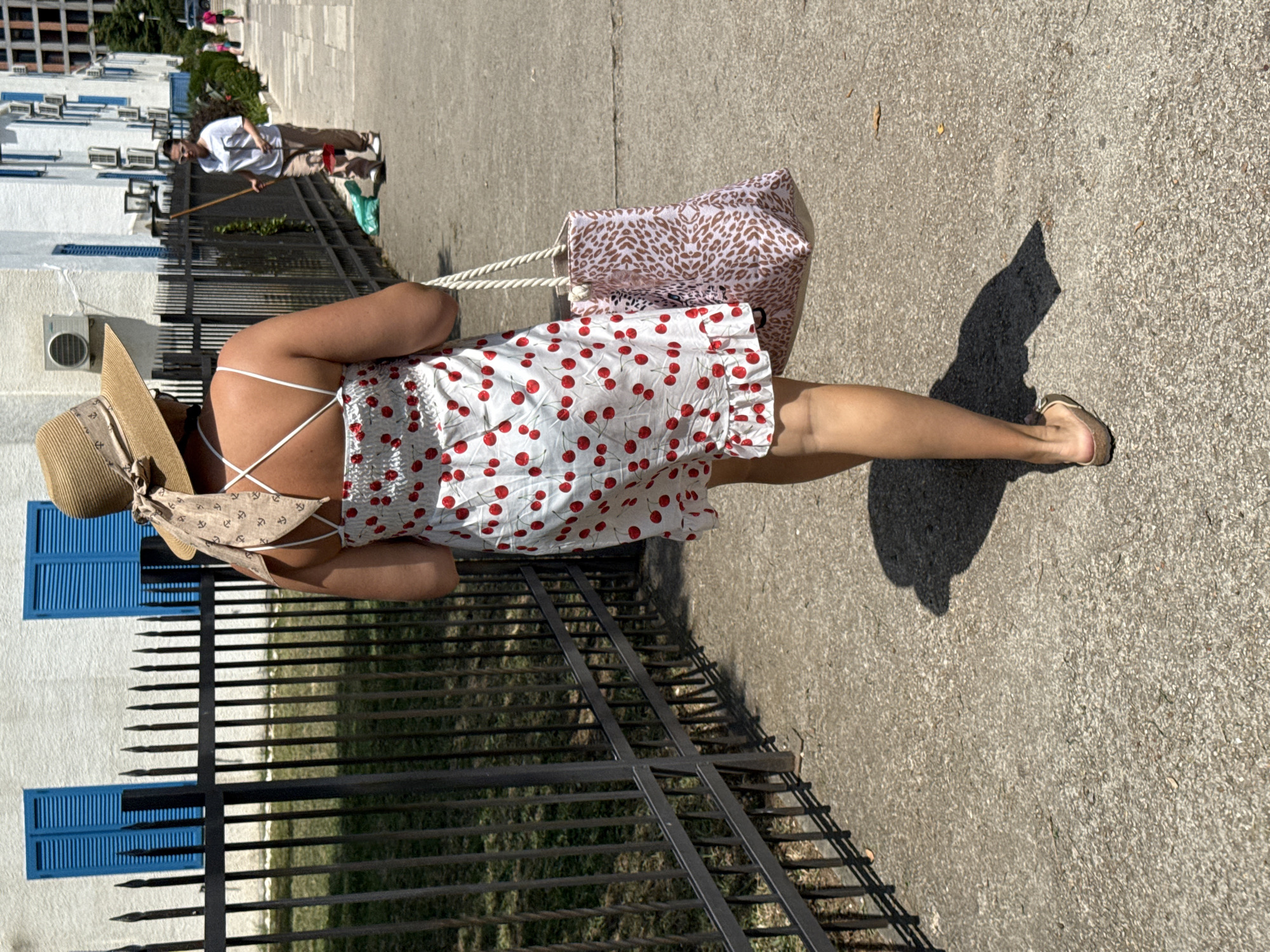 A woman in a white dress with a cherry print and a straw hat walks away from the camera on a paved sidewalk in Boreti, Montenegro. She carries a patterned tote bag on her right shoulder. In the background, a black metal fence separates the sidewalk from a grassy area, and a white building with blue shutters is visible. Another woman, wearing a white t-shirt and beige pants, walks in the opposite direction with a wooden stick. The scene is illuminated by bright sunlight, casting shadows on the ground.