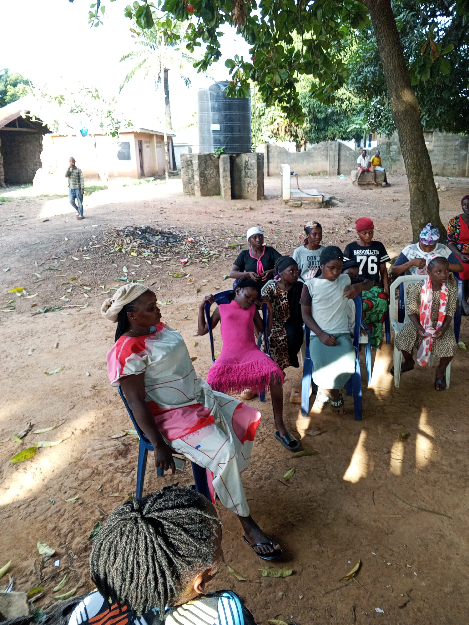 This outdoor image, likely captured in Akwanga, Nigeria, depicts an informal gathering of approximately 15-20 individuals, predominantly women and girls, seated on plastic chairs under the partial shade of a large tree. The setting is a dusty, unpaved area with scattered dry leaves and patches of burnt debris on the ground.
In the immediate foreground, the back of a person with dreadlocks is visible, suggesting the photographer's perspective. Beside them, a woman in a light-colored dress with pink and red patterns and a cream headwrap sits on a blue plastic chair, looking towards the right side of the frame. Behind her, a group of women and girls are arranged in rows on similar chairs, generally facing the right, implying a focal point or speaker outside the visible area. Their attire is casual; one young girl wears a bright pink ruffled dress, and another is in a patterned dress with a white and red scarf. Visible text on clothing includes "BROOKLYN 76" on a black t-shirt and "DON'T QUIT" on a grey t-shirt.
The background shows a semi-rural environment with simple, light-colored buildings, some featuring corrugated metal roofs. A prominent black water tank on a concrete stand is situated in the mid-ground, near a visible water pump and a concrete block wall. Further in the distance, a man walks past a building on the left, and two men are seated on a low concrete structure on the far right.
The scene is illuminated by bright daylight, casting distinct shadows that suggest midday or early afternoon under clear, sunny weather. The overall atmosphere is calm, with subjects appearing attentive or relaxed, indicative of a community meeting, casual discussion, or social gathering.