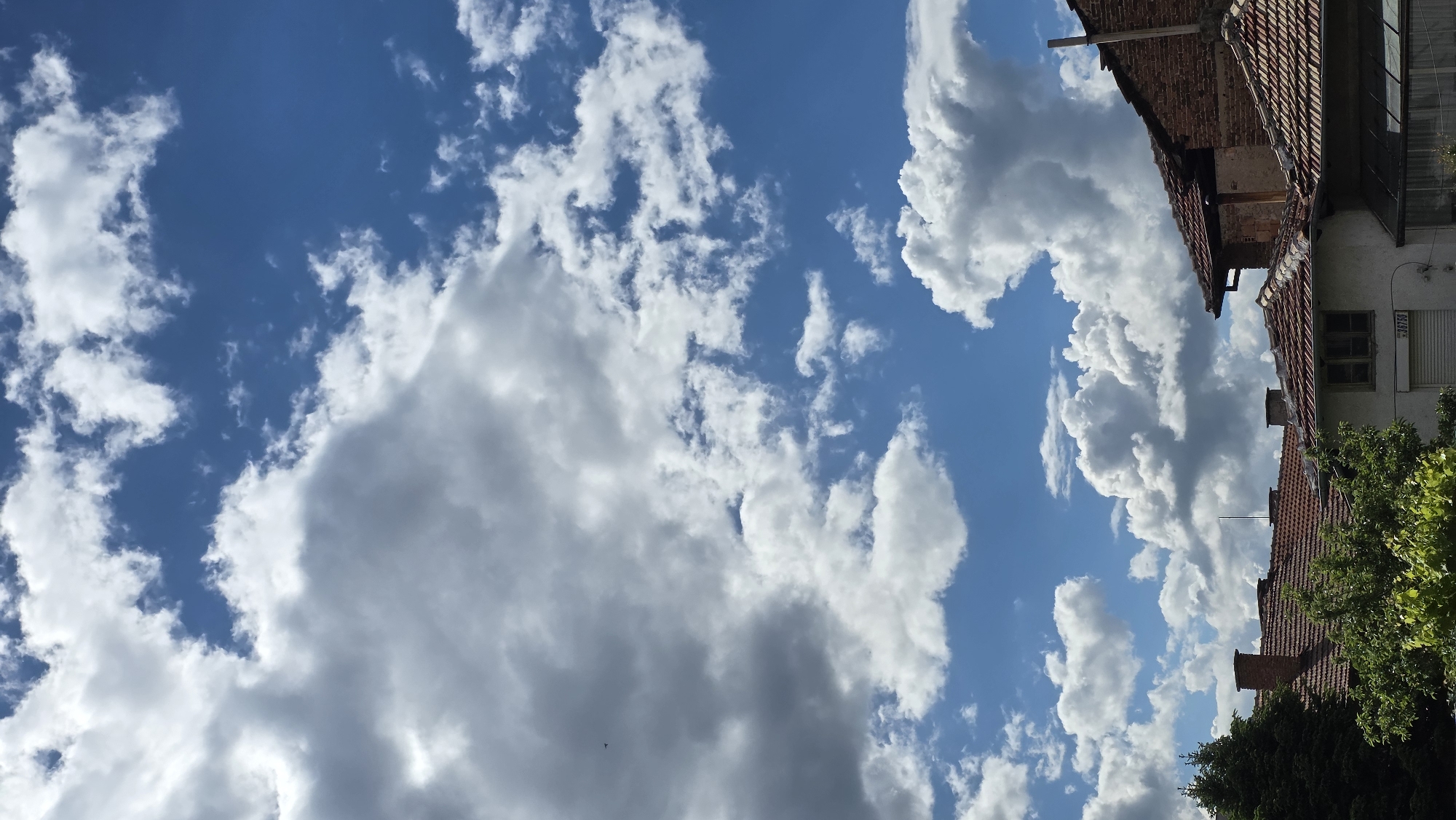 The image depicts a sky with scattered cumulus clouds over Blagoevgrad, Bulgaria. The lower right of the frame shows portions of buildings, including red-tiled roofs, brickwork, and greenery. A section of a white building with a window and a partially rolled-down shutter is also visible. The perspective is upward, emphasizing the sky and clouds.