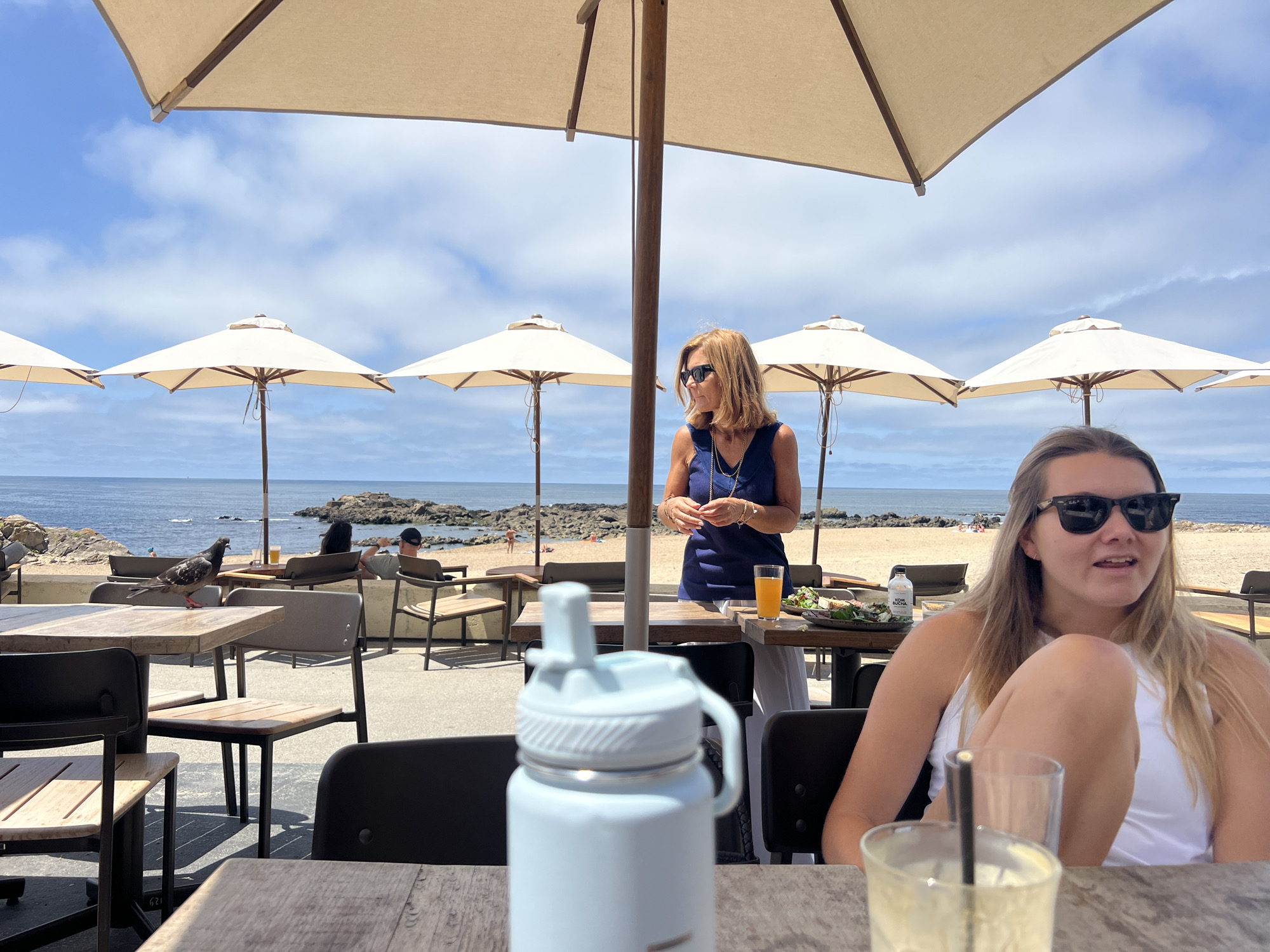 An outdoor cafe with a view of the Atlantic Ocean in Porto, Portugal, features several tables with chairs under beige umbrellas. In the foreground, a light blue water bottle is on a wooden table. To the right, a young woman with blonde hair and sunglasses is seated, looking away from the camera. Behind her, a middle-aged woman in a dark blue dress stands at a table holding a glass of orange juice. A pigeon is perched on a table in the middle ground. The background shows a sandy beach with a few people and the ocean under a partly cloudy sky.
