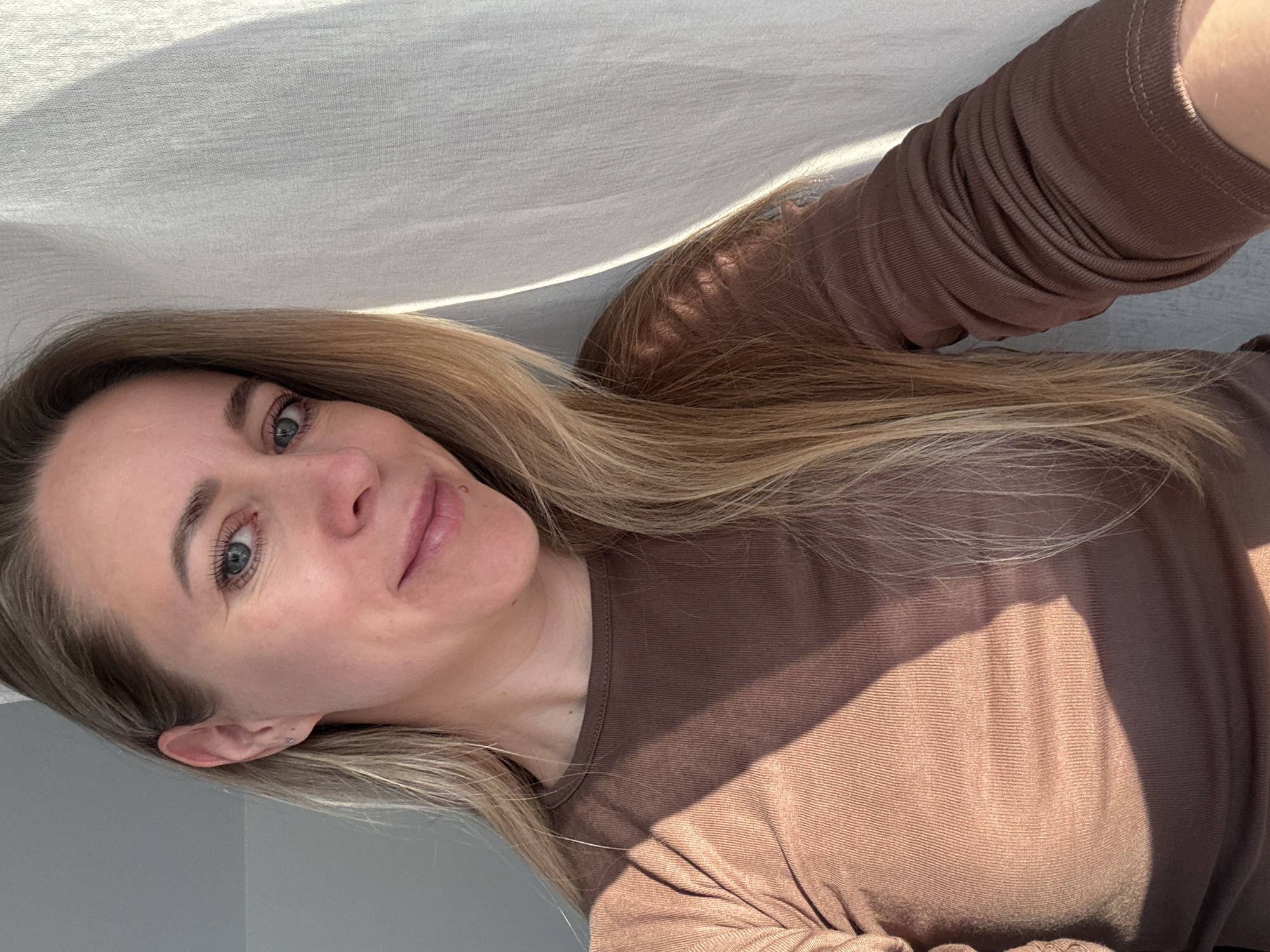 A woman with blonde hair and blue eyes is posing for a selfie indoors. She is wearing a brown long-sleeved shirt. A white cloth is draped above her. The room is lit by natural light, creating shadows on her face and clothing. The background wall is gray. The image was taken somewhere, but not necessarily, in the city of Chaikovsky, Russia. There is no specific evidence to suggest its specific location.