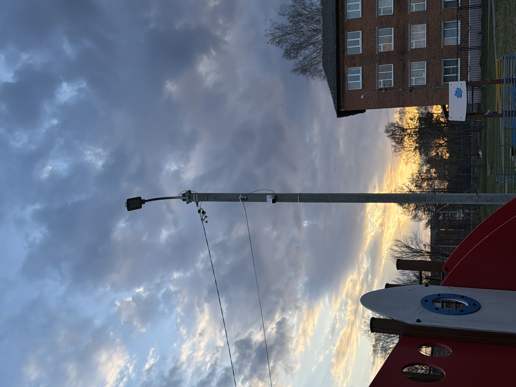 The image shows a playground in Reshetnikovo, Russia. A red and white playground structure is visible in the foreground. A tall grey light pole with wires extends upward, reaching towards a cloudy sky. Behind the light pole, there is a brick building with multiple windows. The sun is setting behind the building and trees, creating a bright golden light. A sign with a blue logo is mounted to the brick building next to a colorful play structure.