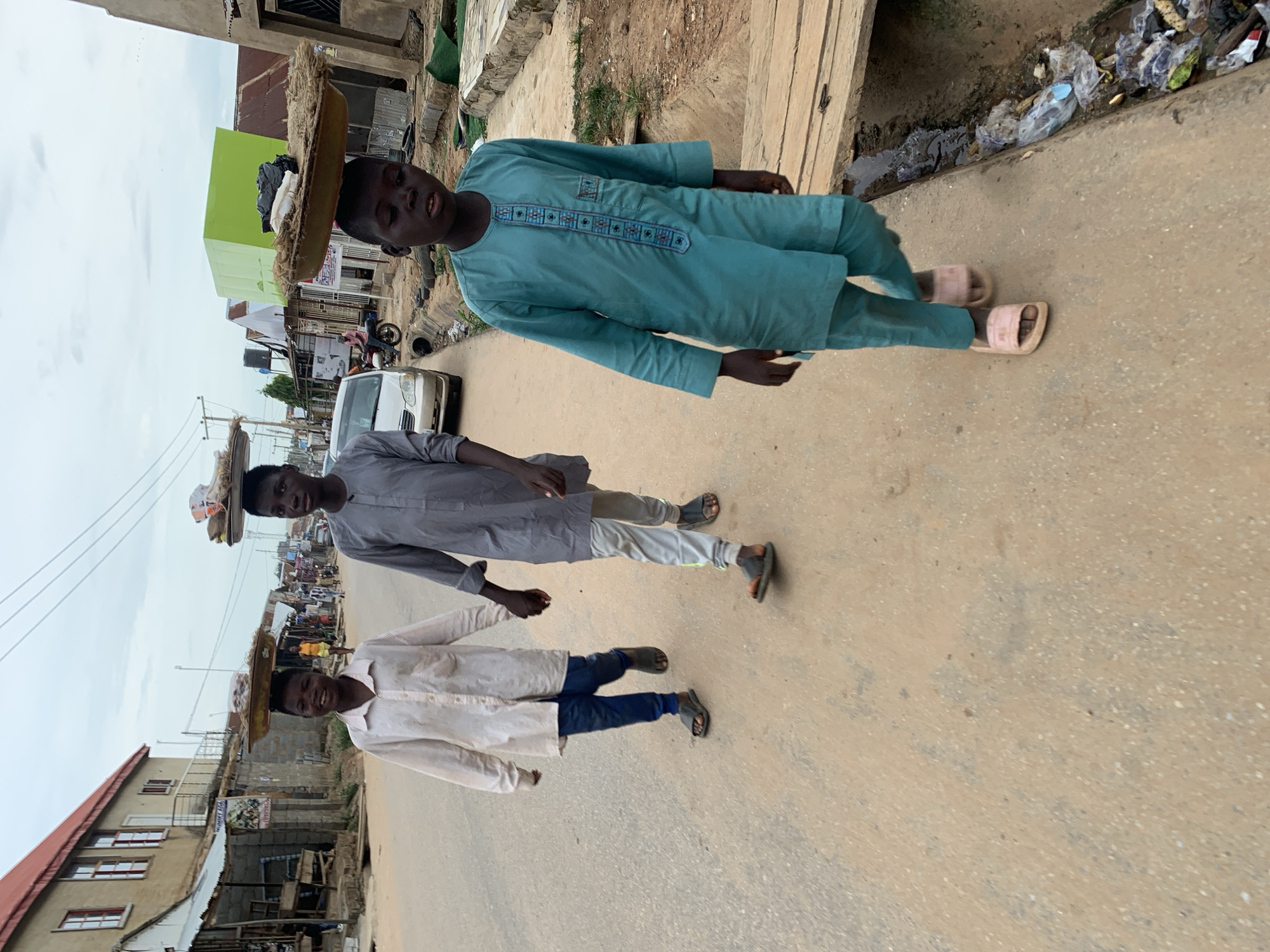 Three young boys walk on a dirt road in Abuja, Nigeria, each balancing a large woven tray of goods on their head. The boy on the left wears a light-colored, long-sleeved shirt and dark pants, with dark sandals. He is holding hands with the middle boy, who is dressed in a gray, long-sleeved tunic and light gray pants, also wearing dark sandals. The boy on the right is in the foreground, wearing a turquoise-colored tunic and pants with pink slides on his feet. He is looking ahead. Behind them, buildings with corrugated metal roofs and a white car are visible. The environment appears to be a commercial or residential area with shops and open stalls lining the road. There is debris and trash in a drainage ditch to the right of the road. The sky is overcast.