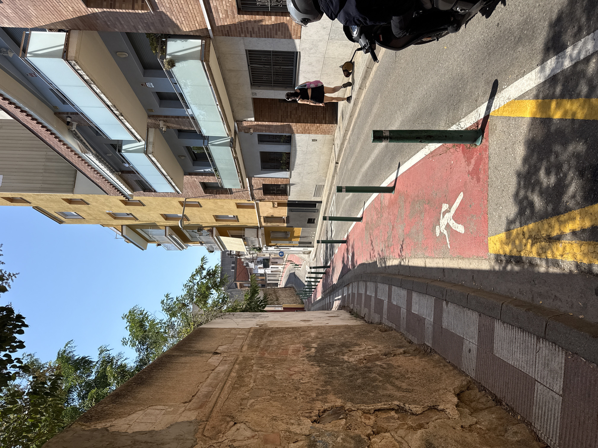 The image depicts a street scene in Blanes, Spain. A narrow, curved street is visible, lined with buildings on the right and a textured, light brown wall on the left. The street has a red-painted lane with a white silhouette of a pedestrian, suggesting a designated walking or cycling path. Green bollards are spaced along the edge of this lane. A woman wearing a black crop top and skirt, carrying a pink bag, walks away from the camera on the sidewalk. In the foreground, the front of a dark motorcycle with a rider wearing a helmet is partially visible on the right side of the street. The sky is clear and blue, and the sunlight casts shadows on the ground. Trees and foliage are visible overhanging the wall on the left.