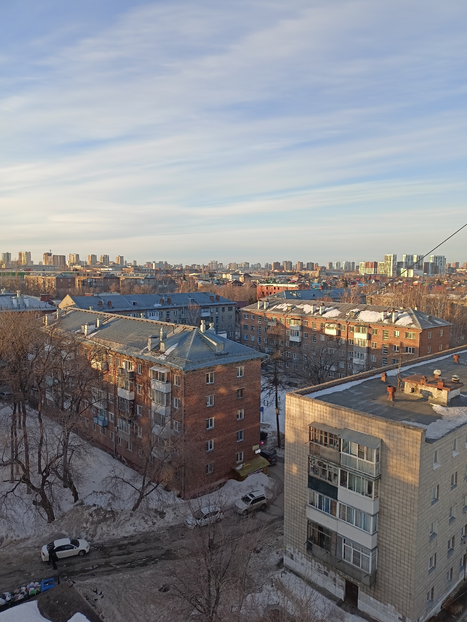 The image shows a cityscape in Novosibirsk, Russia, during winter. Several brick and concrete apartment buildings with snow-covered roofs dominate the foreground. Bare trees dot the landscape, with patches of snow visible on the ground. In the distance, the skyline is filled with more buildings, and the sky is a mix of light blue and wispy clouds. At street level, two cars are visible: one white sedan and another silver SUV type vehicle. A person is standing near the white sedan.