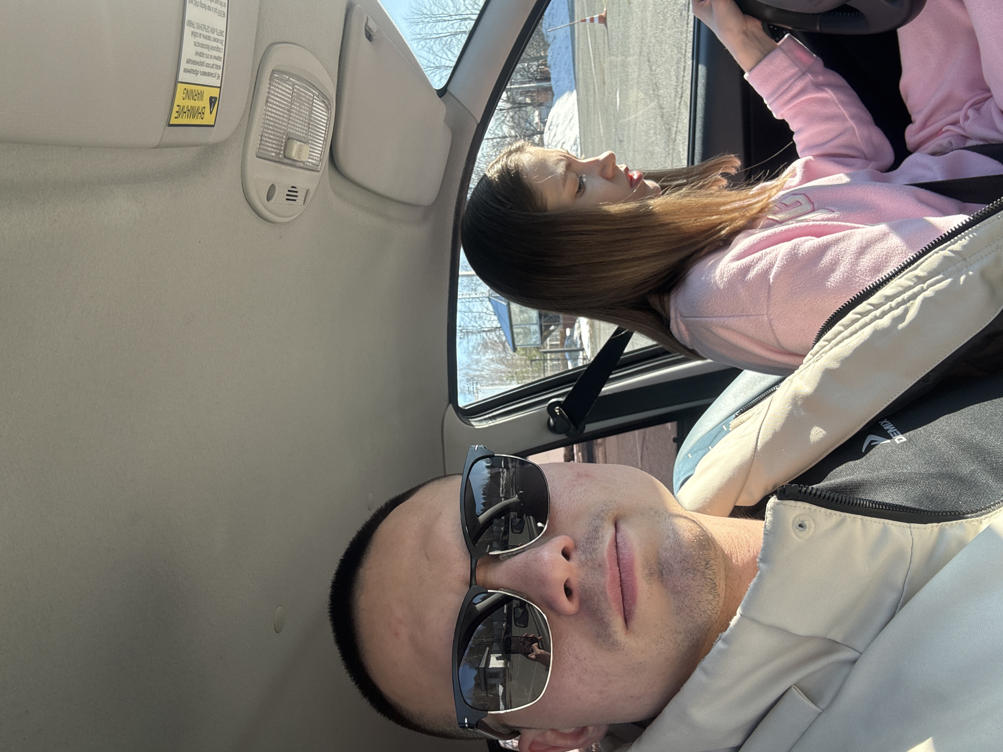 The image is a selfie taken inside a car. A man with sunglasses is in the foreground. Behind him, a woman is visible, presumably driving. The outdoor scenery visible through the car window appears to be a winter landscape. Based on the possible identification on the clothes of the man, the location could be Chaikovsky, Russia.
