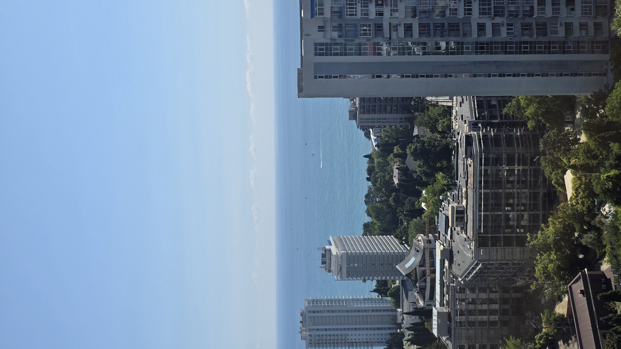 The image captures a coastal city skyline under a clear blue sky. In the foreground, several modern buildings, including high-rise apartment complexes and a partially constructed building with scaffolding, dominate the scene. These buildings are interspersed with lush green trees. Further back, the vibrant blue sea stretches to the horizon, dotted with a few small boats. The architecture suggests a contemporary urban development near a recreational waterfront. The absence of any visible people or specific identifiable landmarks makes it challenging to pinpoint the exact location within Sochi, Russia, beyond it being a seaside urban area.