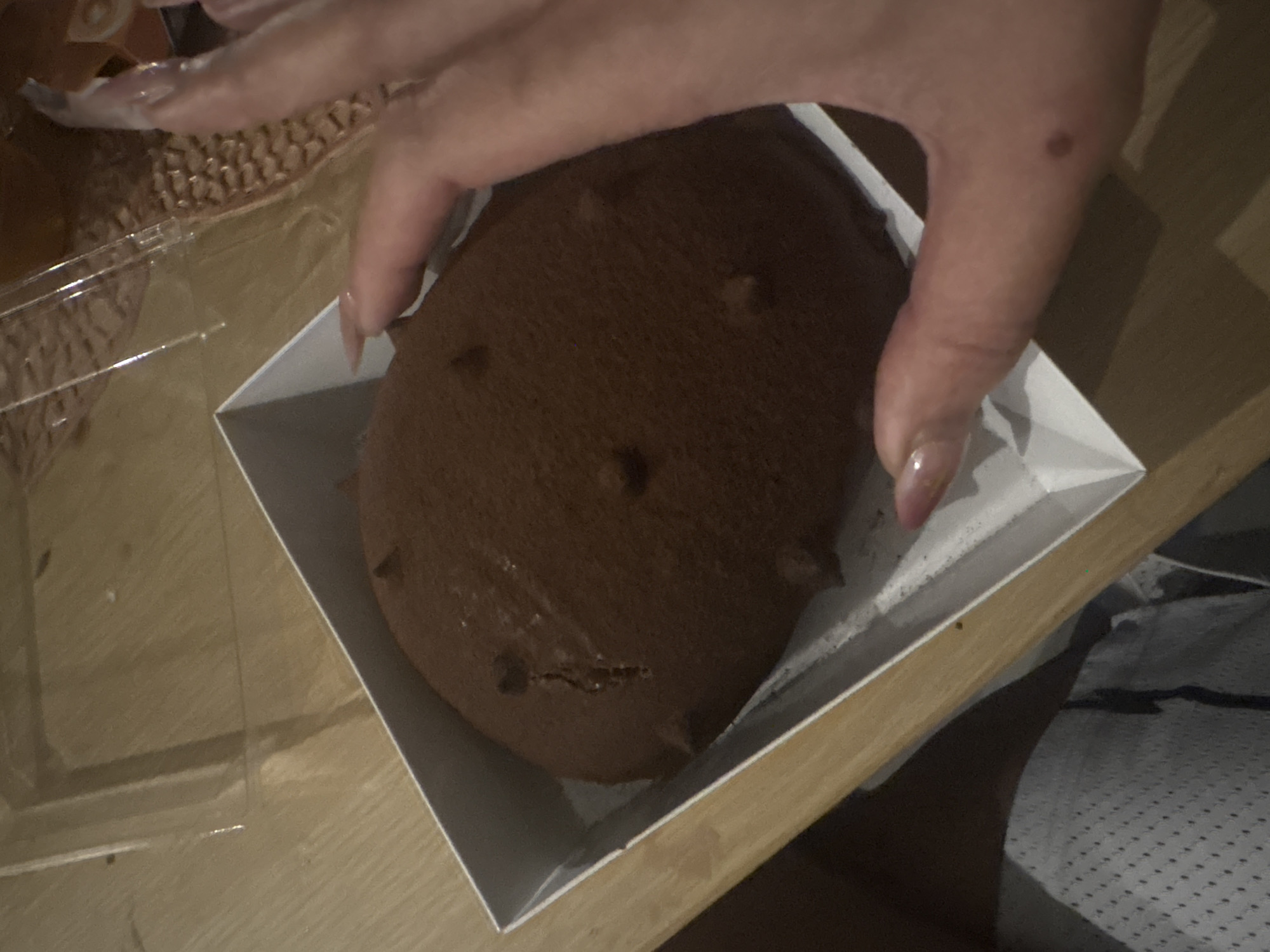 A person's hand is partially visible over a brown, oval-shaped pastry in a white, square container. The pastry is textured with small, raised dots or protrusions. The container is situated on a wooden surface, with a partially visible clear plastic container to the left. The person's nails are long and manicured with a clear, glossy finish. The lower right corner of the image shows a patterned fabric or clothing. The location in the city of Unknown, Unknown is not discernible.