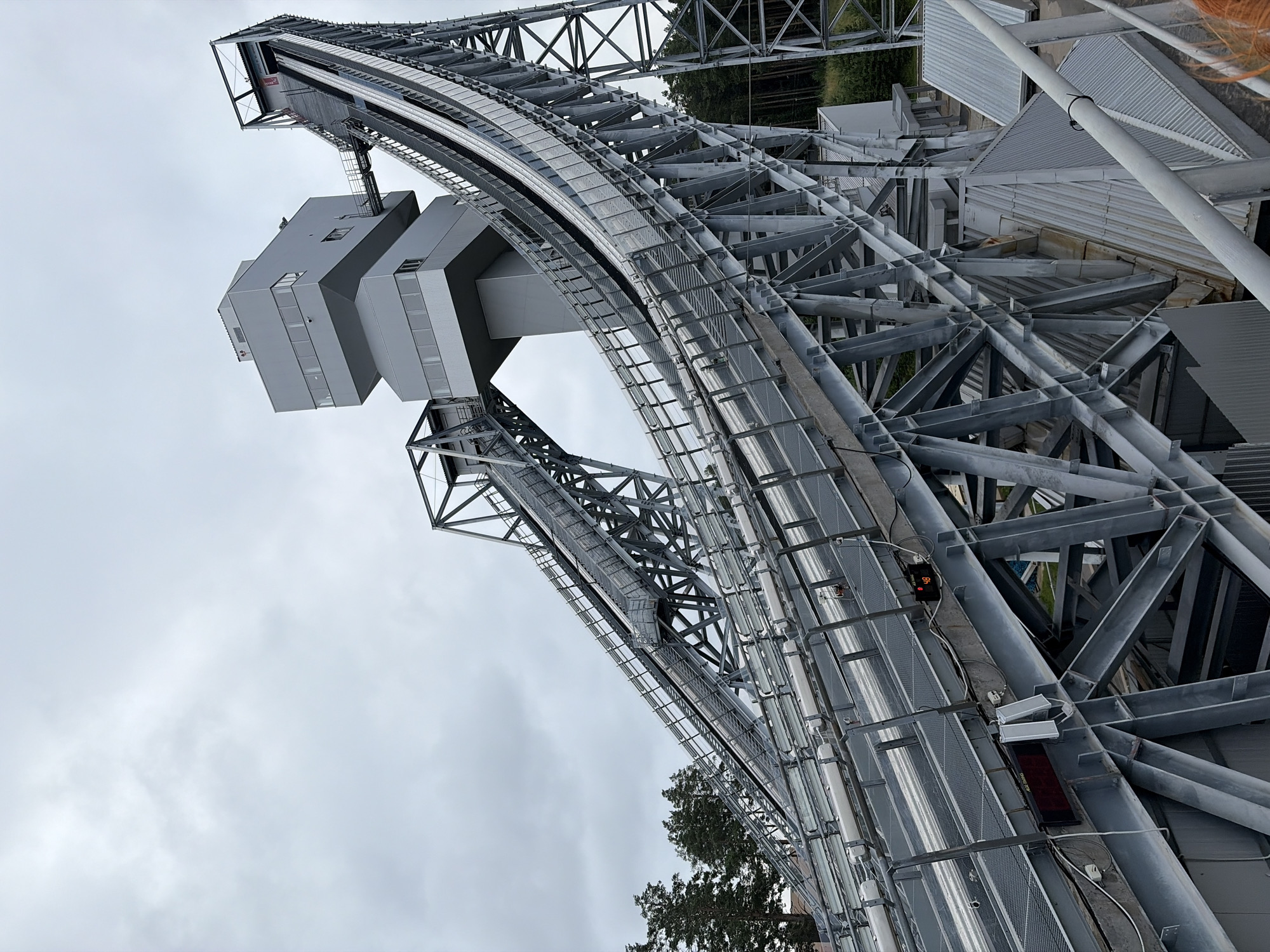 The image shows a ski jumping facility in an overcast outdoor environment. The main subject is a large, complex metal structure of a ski jump, with two distinct inclined tracks leading up to a higher platform. The structure is made of gray metal beams, supports, and railings. Along the sides of the tracks, there are rows of lights and some electrical equipment, including a digital display showing "48" and a red LED sign. A modern, multi-story gray building with windows is situated at the top of the structure, connected by a walkway. Trees are visible in the background on the right side of the image, suggesting a wooded area. The sky is cloudy and gray. There are no people visible in the image, and no actions or interactions are depicted. The location is identified as the city of Perm (Прикамский is an area within Perm), Russia.