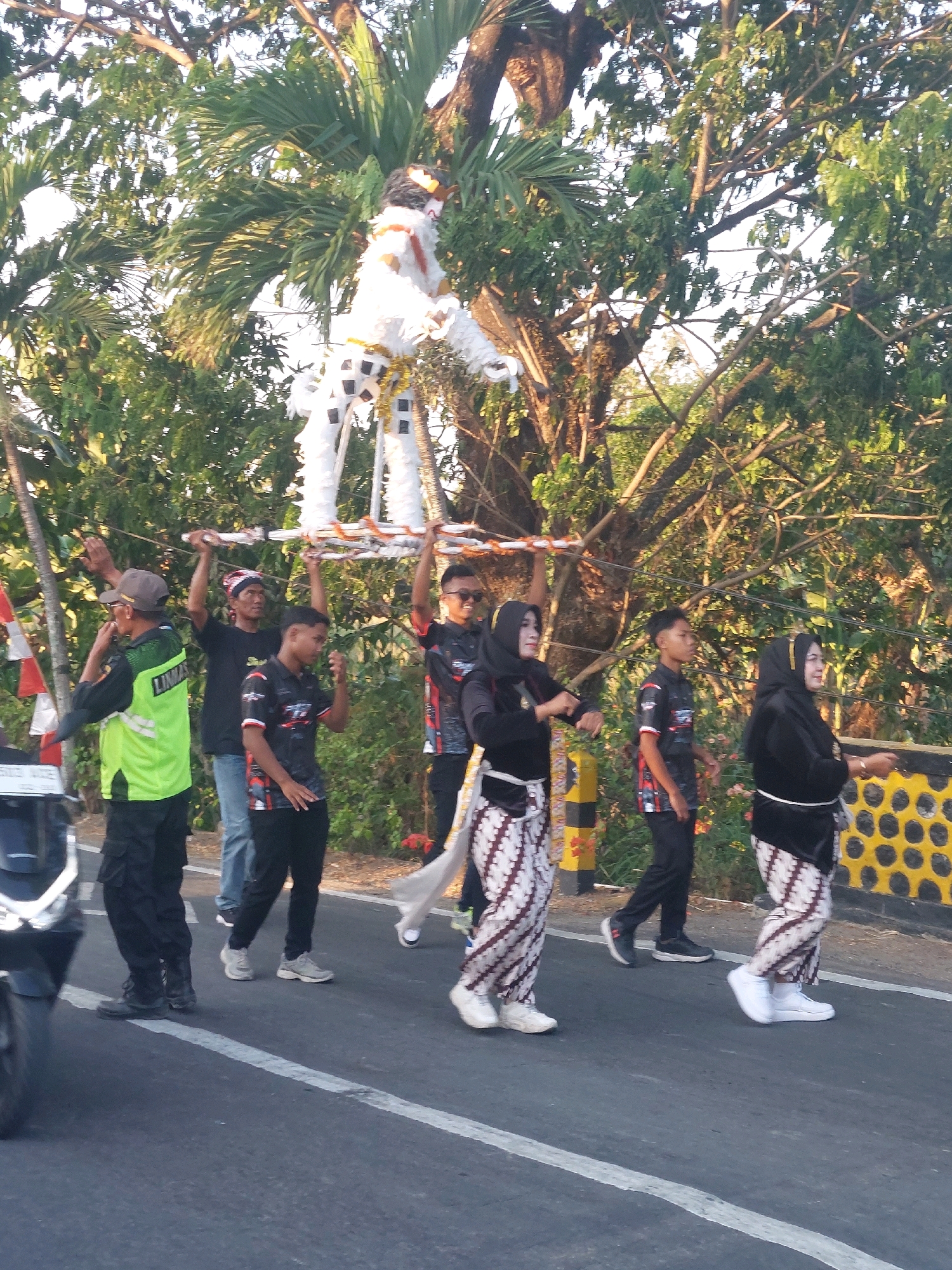 In Jono, Indonesia, a group of people are participating in a procession along a paved road. The central focus is a large, white, horse-like effigy adorned with orange and black accents, carried aloft on a platform by four individuals. The effigy has a black mane and tail, and its body is made of a white, textured material, possibly paper or fabric.
To the left of the effigy carriers, a man in a bright green vest with the word "LINMAS" visible on the back and a grey cap is walking and gesturing with his left hand. Behind him, another man in a black shirt and blue jeans is also participating in the procession.
To the right of the effigy carriers, a group of individuals are walking in unison. Two women are dressed in black headscarves and batik patterned skirts, and are walking with their arms held out to their sides. A young man in a black shirt with red and white accents and black pants is walking between them. Further to the right, another individual, partially obscured, is also dressed similarly to the women.
The environment includes a road with a white line marking its edge, bordered by a low wall with circular yellow and black decorative elements. Lush green trees with prominent branches and foliage line the background, suggesting an outdoor setting under daylight. The lighting indicates it may be late afternoon or early morning, with the sun casting warm tones. A motorcycle is partially visible on the far left of the image.