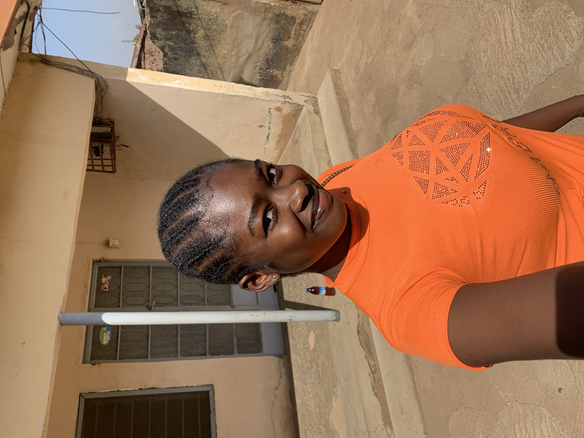 A young Black woman in an orange shirt is smiling toward the viewer. She has her hair braided back. She is standing outside in what appears to be a courtyard area of a residential building. There is a door to a building behind her. The location is Rafin Par, Nigeria.
