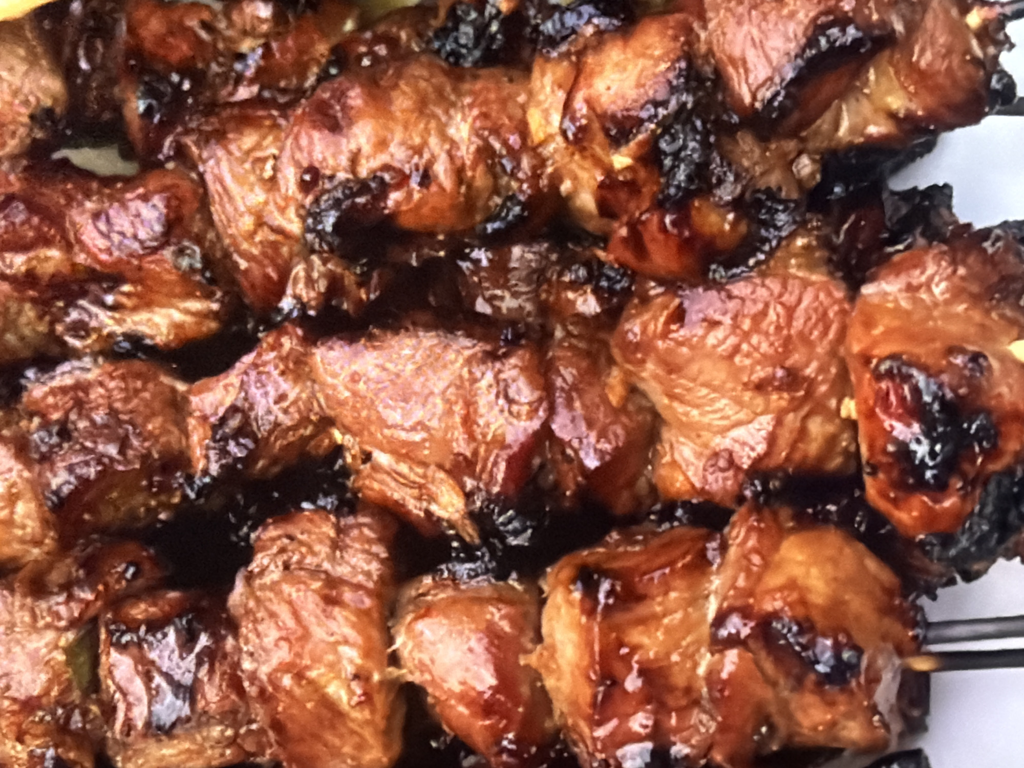 A close-up, overhead shot shows a skewer of grilled meat. The meat appears to be chunks of pork or chicken, glazed with a dark, savory sauce and slightly charred in places. The meat is glistening, suggesting it is hot and freshly cooked. The background is blurred and out of focus, but appears to be a white surface. The image is well-lit, with the light source coming from above and slightly to the left, highlighting the textures and details of the meat.