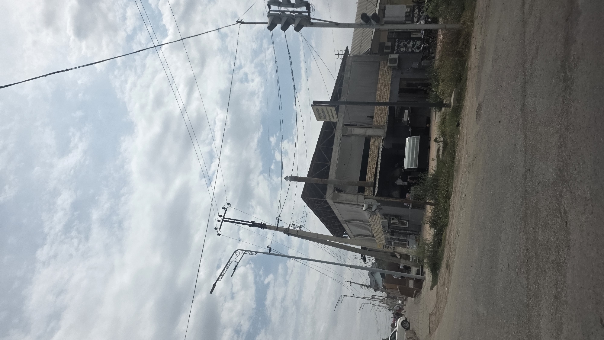 The image shows a street scene in Telimana, Kyrgyzstan, with a building that appears to be a restaurant or cafe on the right side of the frame. The building has a modern design with large glass windows and an outdoor seating area. A traffic light and a street sign are visible next to the building. The street is paved, and there are power lines and utility poles running along the side of the road. A few cars are parked on the street, and some people can be seen walking in the background. The sky is partly cloudy.
