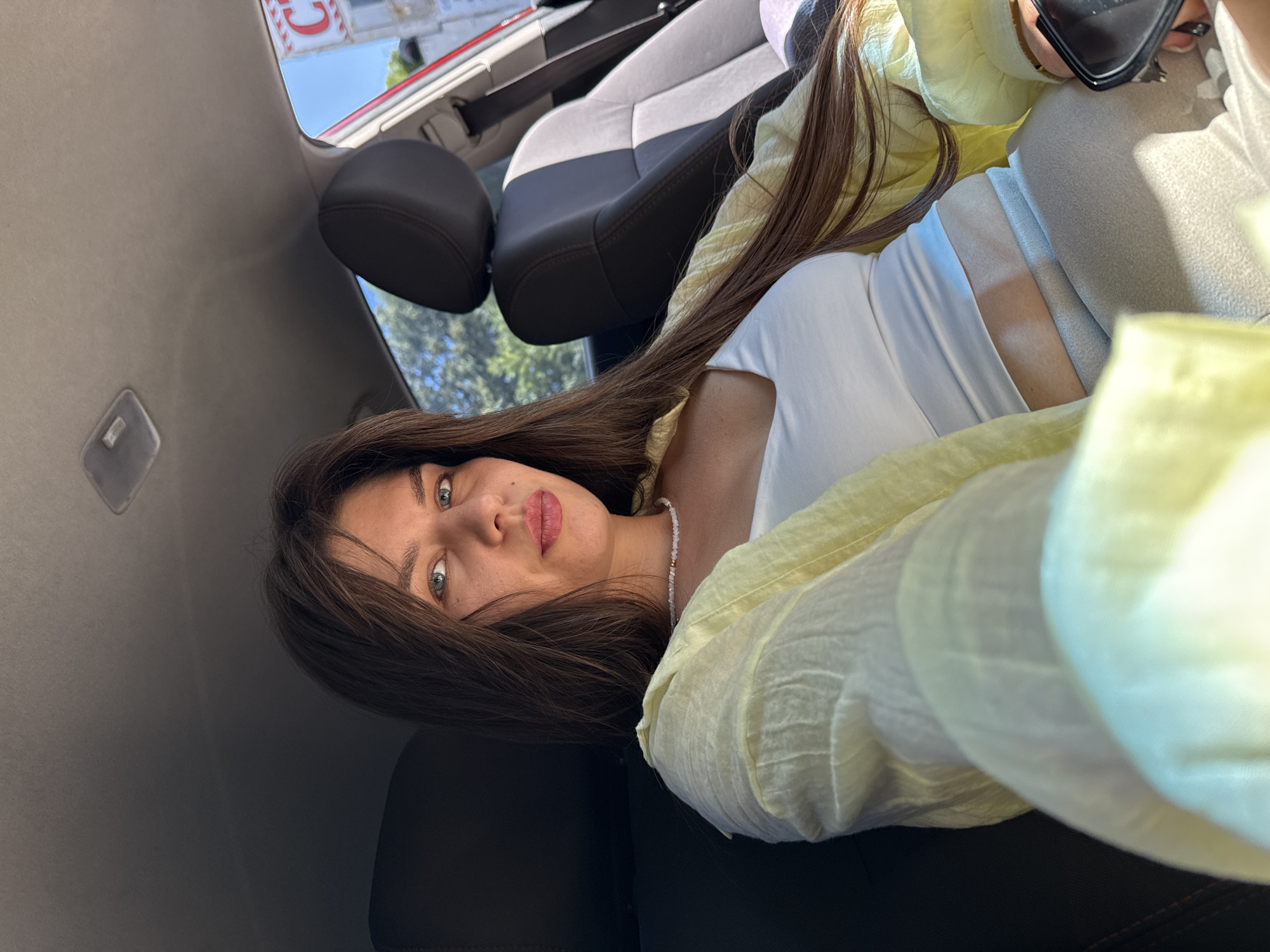 A young woman with long brown hair and blue eyes is seated in the passenger seat of a car. She is wearing a light yellow, oversized shirt over a white tank top and light-colored pants. She is looking directly at the camera, her makeup including pink lipstick. A delicate beaded necklace adorns her neck. The interior of the car is visible, including the dark seats and the headliner. Sunlight streams into the car, casting shadows on her face and clothing. Outside the car, trees and a sign with the letter "C" are visible. The scene appears to be a candid shot, possibly a selfie, taken during the daytime.