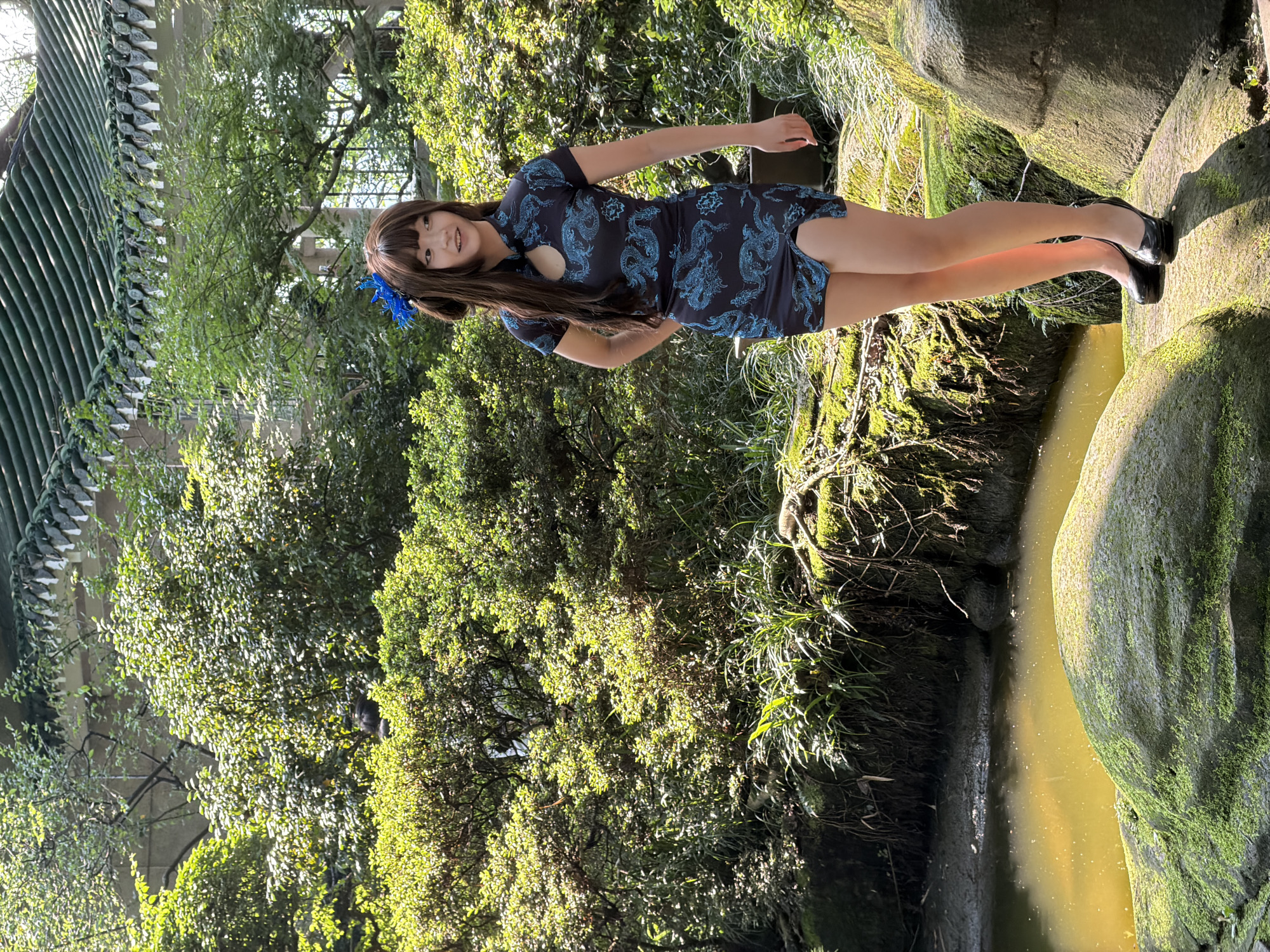 In a lush, sun-dappled garden with moss-covered rocks and a tranquil pond, a young East Asian woman stands facing right, her body angled towards the camera. She is wearing a short, dark dress adorned with a blue dragon pattern, featuring a high collar, a cut-out at the chest, and a slit on the left side. Her dark hair is styled with a blue hair accessory, and she wears black high-heeled shoes. The background features dense green foliage, a traditional Chinese building with a green tiled roof, and sunlight filtering through the leaves. The setting appears to be a serene park or garden, potentially within the Jiangbeicheng Street area of China.