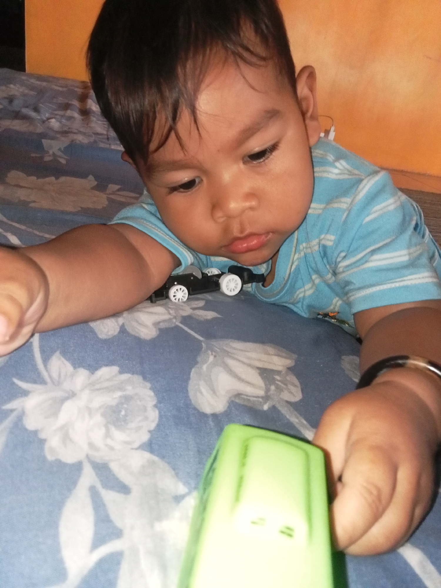 A dark-skinned infant with short, dark hair lies on their stomach on a blue and white floral patterned surface. The infant wears a light blue horizontally striped t-shirt. Their head is turned to the right, and their gaze is directed downwards. In front of the infant, to the left, is a small black toy car with white wheels. To the right, the infant's right arm is extended forward, and their hand is reaching for a bright green toy. A thin black bracelet is visible on the infant's right wrist. The background consists of an orange wall and the blue and white patterned surface. The setting appears to be a domestic environment. The location is identified as Pancurwening, Indonesia.