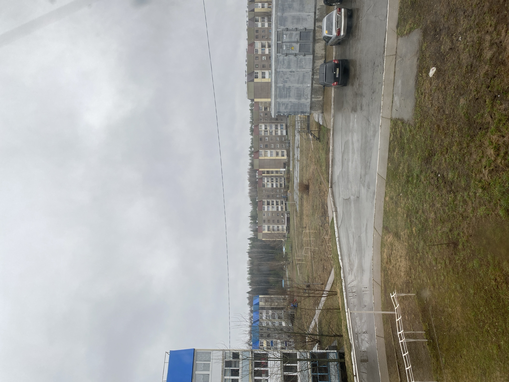 This image, when viewed in its correct landscape orientation, presents an overcast daytime scene of a residential area in Markovskii, Russia.
The sky is uniformly grey, indicating heavy cloud cover or a dull, damp day. In the mid-ground and background, multiple multi-story apartment buildings are visible. These structures feature facades in beige, grey, and light yellow tones, with numerous windows. Some buildings, particularly in the closer view, have distinctive blue roof sections or coverings, which might suggest ongoing construction, renovation, or specific architectural features. Behind a cluster of these buildings, a dense line of bare deciduous trees stretches across the horizon, characteristic of late autumn, winter, or early spring.
The ground consists of a network of wet, dark asphalt roads and pathways, interspersed with areas of sparse, brownish-green grass and bare earth. The dampness of the surfaces suggests recent rain or generally wet conditions. A large, grey, industrial-style container or utility box with a yellow warning triangle is situated next to one of the apartment buildings. Several metal structures, resembling old playground equipment or simple fences, are scattered across the grassy plots.
Multiple cars are parked along the wet asphalt roads. A silver sedan, possibly a Lada Granta, has a visible Russian license plate that appears to be "669 MM 199". Another black sedan, likely also a Lada model, clearly displays the license plate "450 ОВ 790". There are no people visible, and no discernible activity is taking place, contributing to the quiet, still atmosphere of the scene. The overall impression is that of a typical urban residential area on a cool, overcast day.
