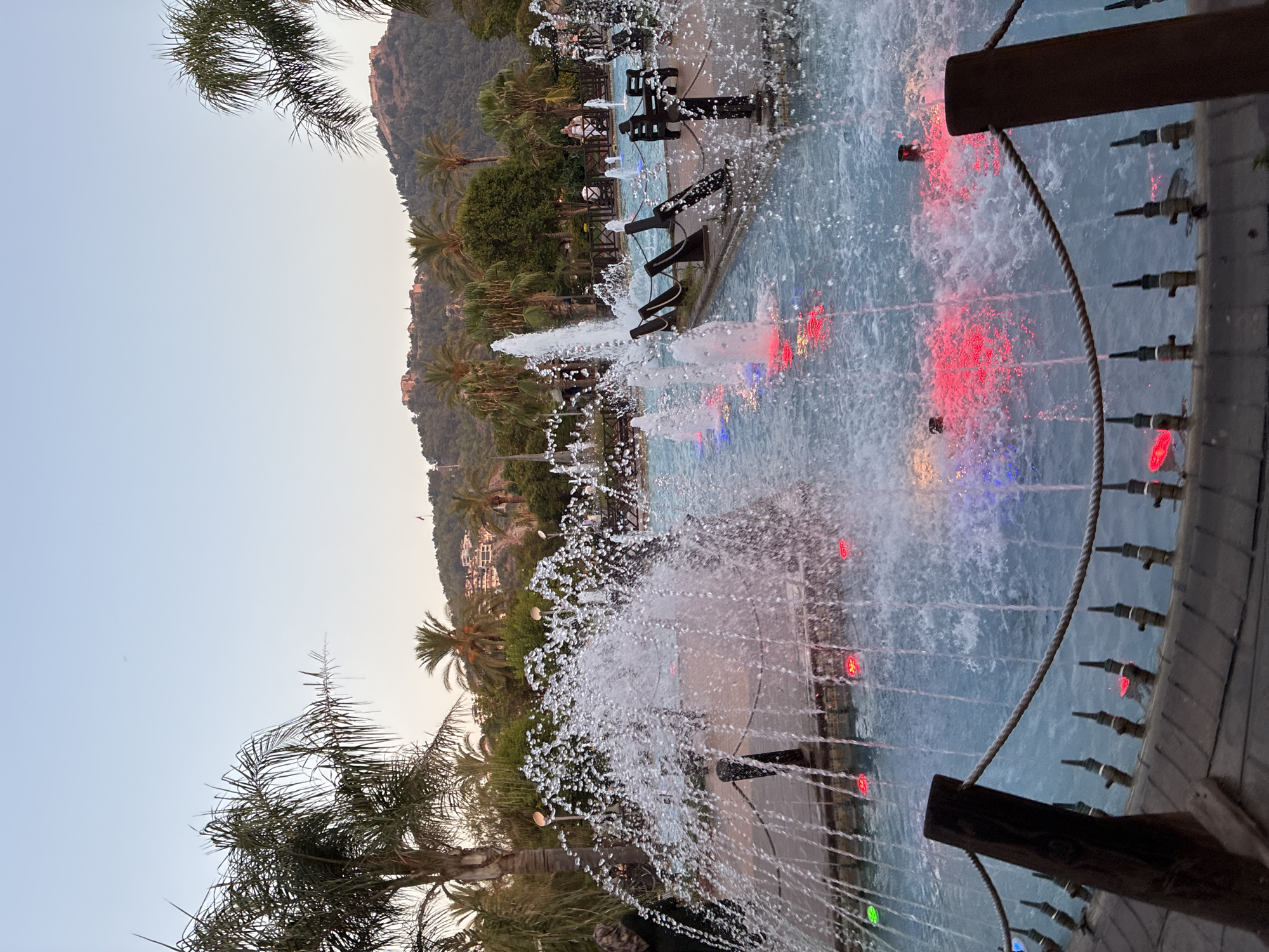 The image depicts a public fountain area in Alanya, Turkey, as suggested by the presence of palm trees and a mountainous backdrop with visible structures, possibly historical fortifications. The fountain features multiple jets of water creating a dynamic spray, illuminated by colorful underwater lights, predominantly red and blue, reflecting off the water's surface. The water is contained within a pool with a light blue hue. Surrounding the pool, there are wooden deck platforms with rope railings and some seating areas with dark wooden benches. In the background, lush green trees and palm trees are visible, with buildings cascading down a hillside under a clear, pale sky, indicative of twilight or early evening. The overall scene conveys a tranquil, recreational atmosphere in a coastal city.