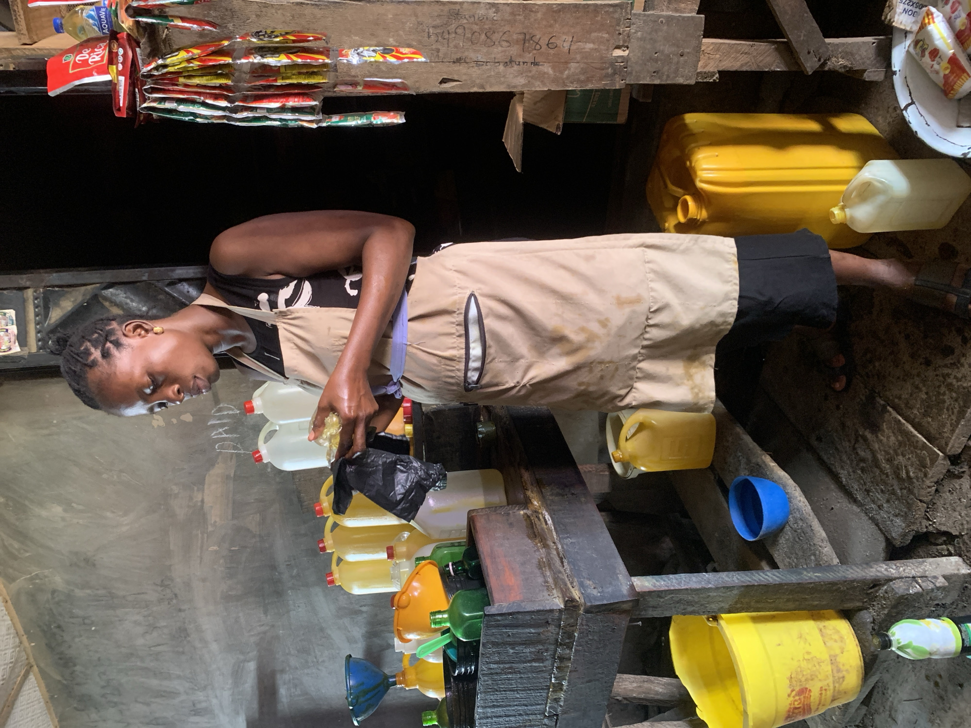 A woman is filling jerrycans with liquid, presumably palm oil, inside a stall in Shagamu, Nigeria. The stall's wooden exterior displays a phone number and a name. The woman wears an apron and is surrounded by plastic containers, funnels, and jugs in various colors. Condiments and beverages are displayed above the stall.