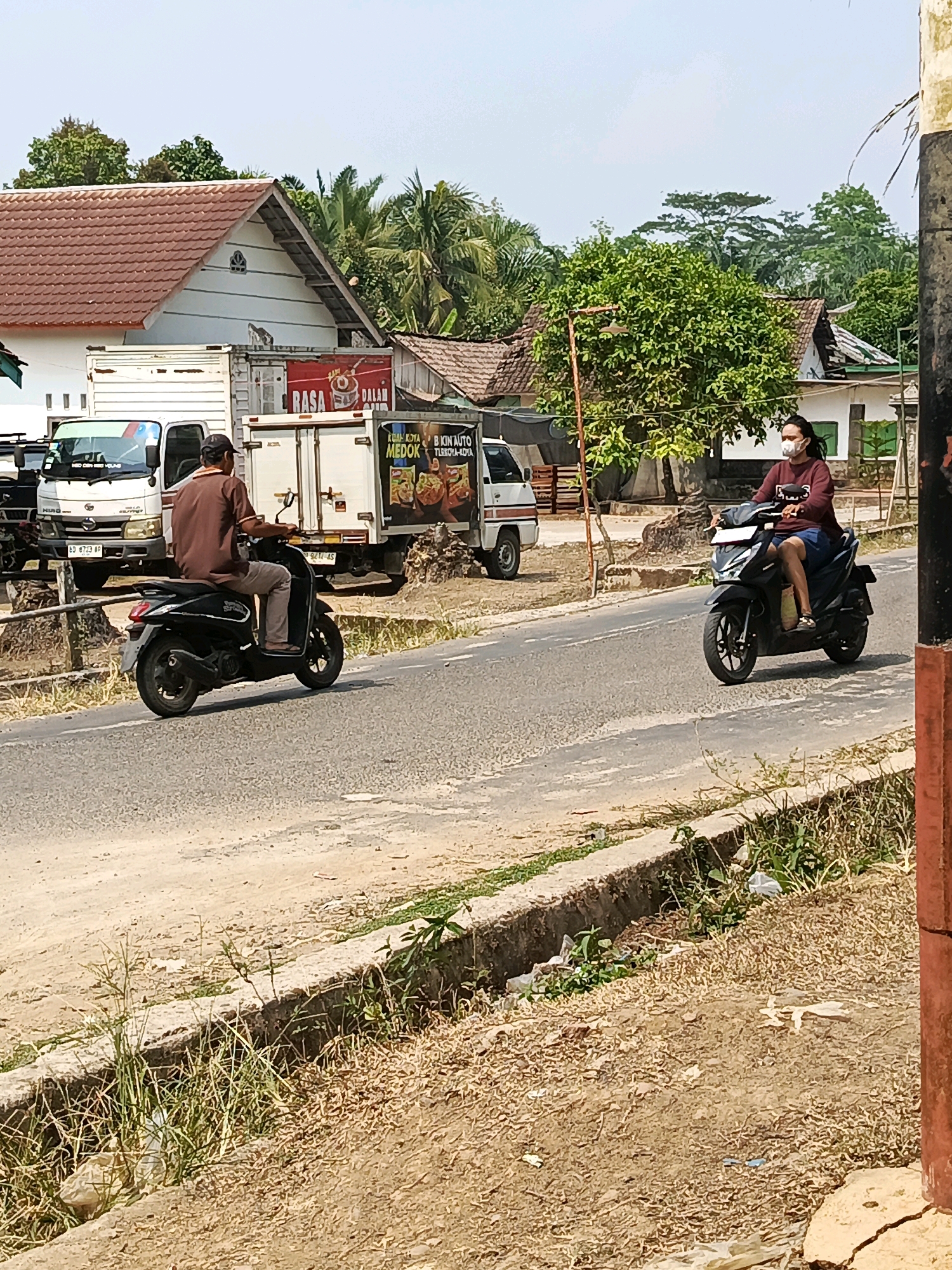 A mid-range shot shows a street scene in Wonosari, Indonesia, with two people on motorcycles. On the left, a man wearing a brown shirt and cap rides a black scooter. To his right, further down the road, a person with long dark hair wearing a maroon sweater and blue shorts, and a face mask, rides a black motorcycle. Both are traveling on a paved road with a drainage ditch on the right side of the frame. Behind the motorcycles, a white truck with a red advertisement panel is parked, and other buildings and trees are visible in the background. The ground is covered with dry grass and some litter. The sky is clear and bright.