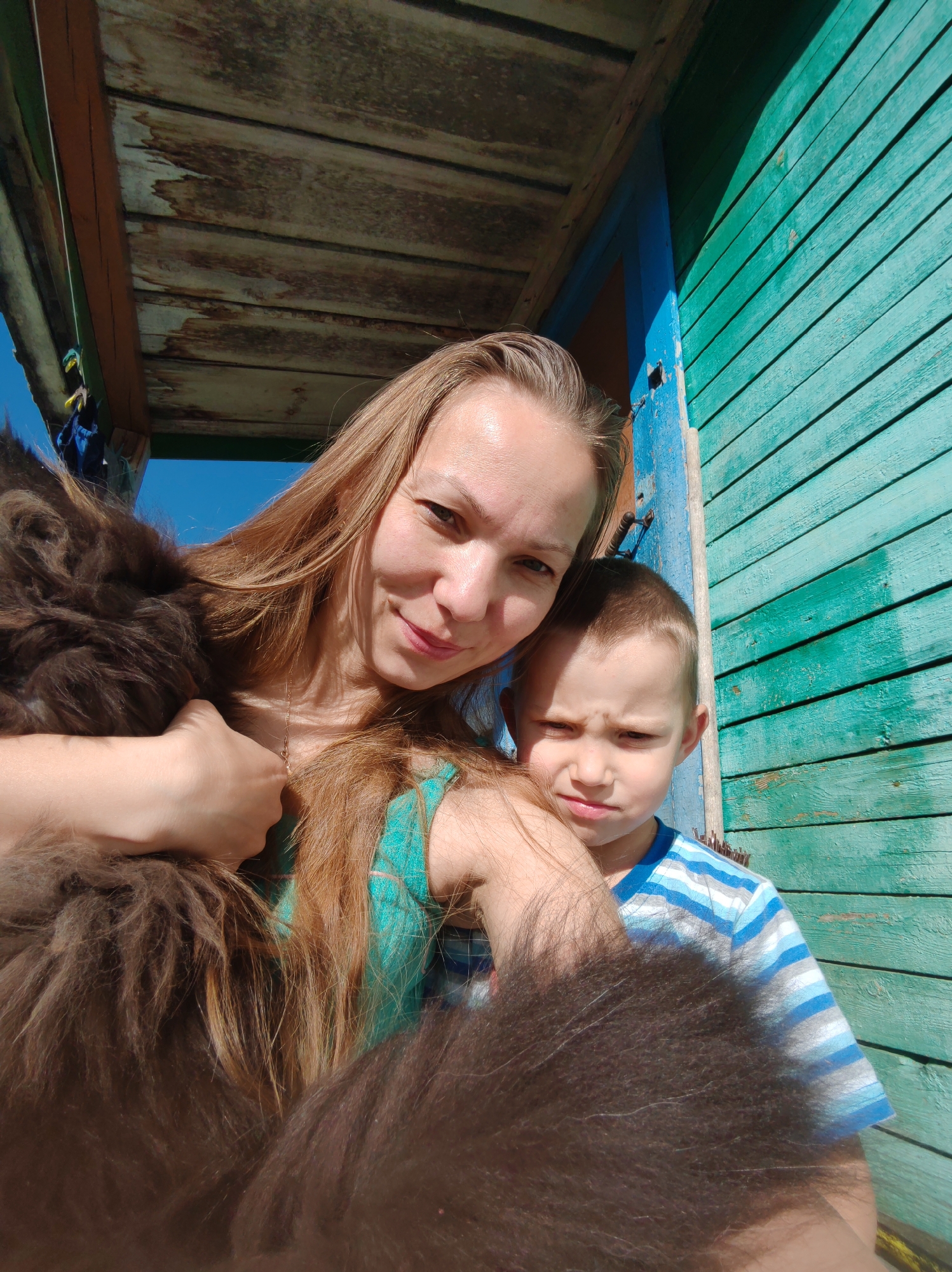 A woman and a young boy are taking a selfie outdoors. The woman is on the left, holding a large, dark, furry animal, possibly a dog, and looking at the camera with a smile. The boy is on the right, looking at the camera with a slightly furrowed brow. They are standing in front of a turquoise-colored wooden building with a blue door frame. Above them is a rustic wooden structure, possibly a porch or an overhang, with weathered planks. The sky is visible and appears to be blue. The overall impression is a casual, everyday scene likely taken at a residential property.