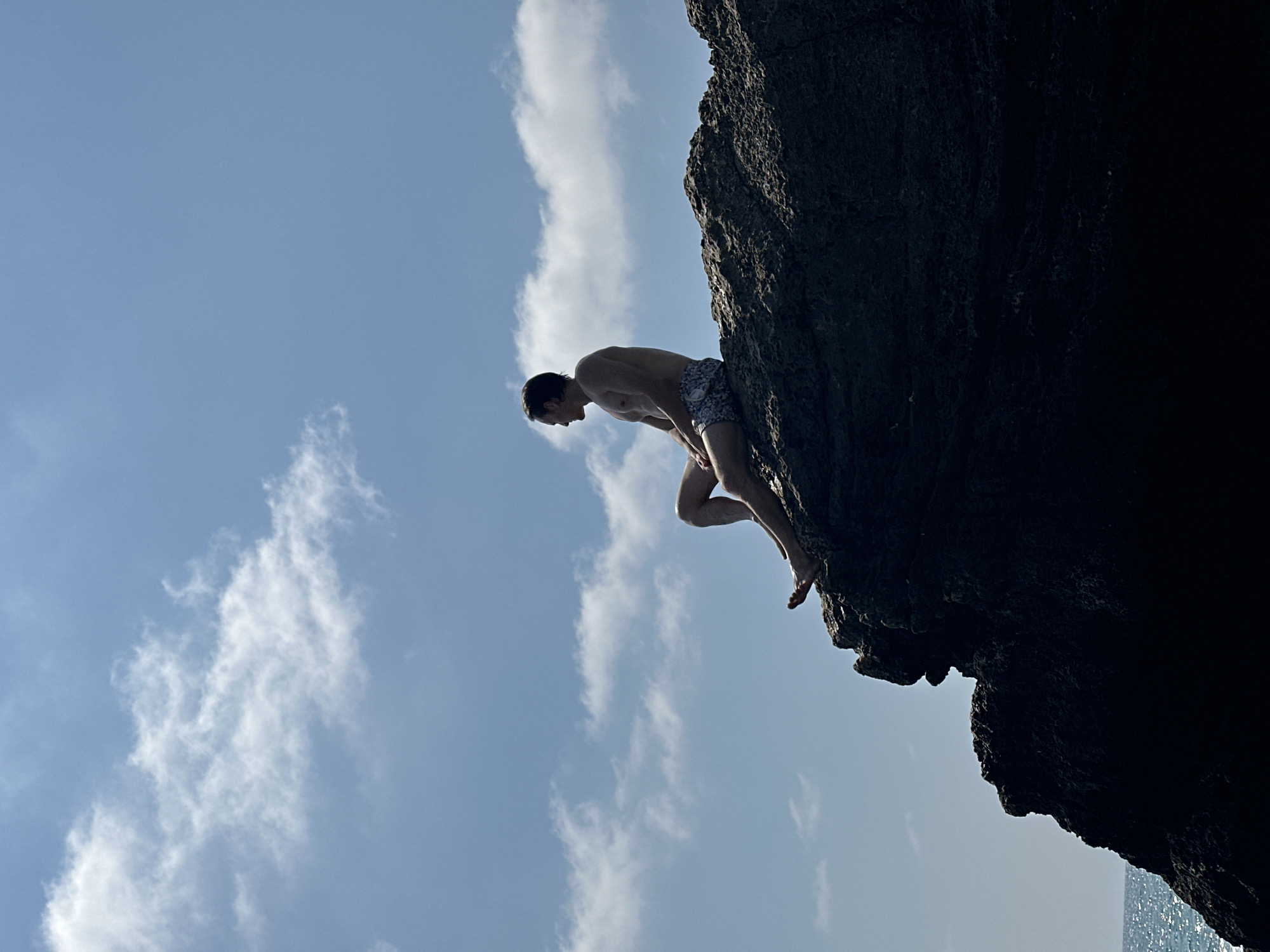 A silhouette of a shirtless man wearing patterned swim trunks sits on the edge of a dark, craggy rock formation. He is looking out to his left, with the bright, cloudy sky and a distant ocean in the background. The rock formation is positioned in the sea, with glistening waves visible below and to the left of the frame. The overall scene suggests a coastal location in Hof HaCarmel Regional Council, Israel, with the man enjoying a view of the Mediterranean Sea.
