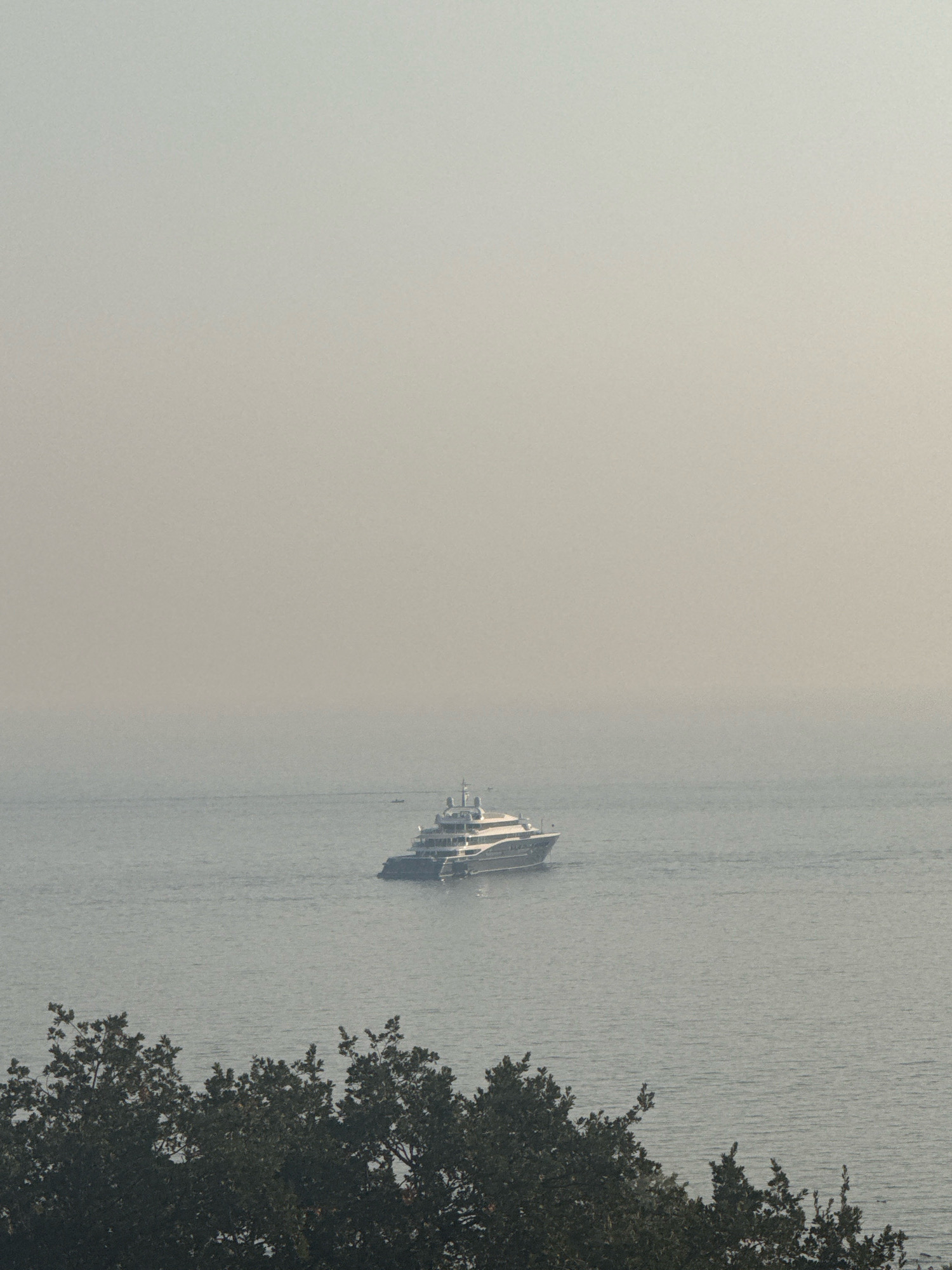The image shows a wide shot of the Boka Kotorska Bay near Boreti, Montenegro, under a hazy, overcast sky. In the middle distance, a large, multi-decked white yacht is anchored on the calm, grey water. The yacht has a sleek, modern design with multiple levels and visible windows. In the foreground, the dark green foliage of trees lines the bottom of the frame, suggesting an elevated viewpoint overlooking the bay. The overall atmosphere is muted and tranquil, with a sense of atmospheric haze obscuring the horizon. There are no people visible in the image.