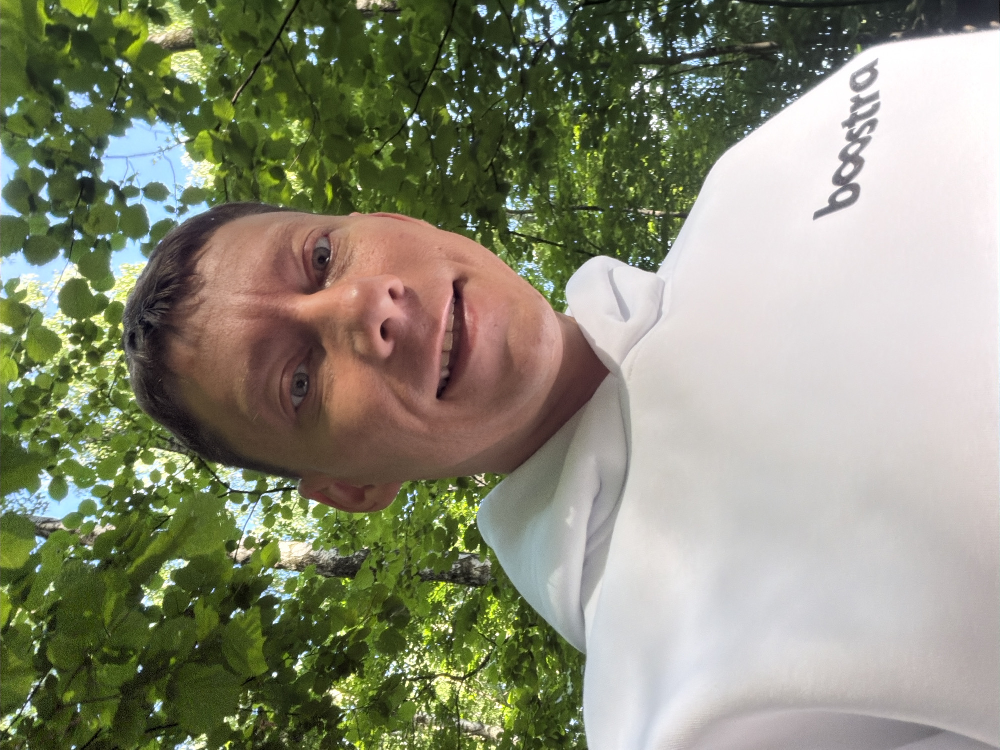 A close-up, self-taken photograph shows a fair-skinned adult male with short brown hair, wearing a white hooded sweatshirt. The sweatshirt has the word "boostra" printed in black on the right chest area. The man is outdoors, with a backdrop of lush green leaves and dappled sunlight. The foliage suggests a wooded or park-like setting, possibly in Moscow, Russia, though no specific landmarks are visible. The man is smiling, and the camera is positioned at a slightly low angle, looking up at him. The sky is visible through the leaves, indicating it is daytime.