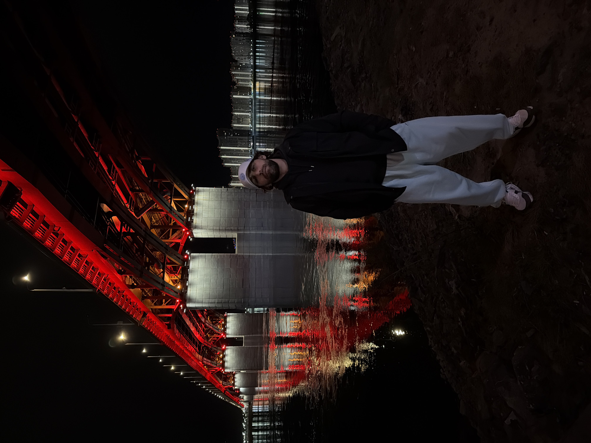 A low-angle, nighttime shot features a man standing next to the Yenisey River and the Vinogradovsky Bridge in Krasnoyarsk, Russia. The man, facing the camera, wears a white hat, dark jacket, and light-colored sweatpants. He is positioned on a dark, rough terrain near the water's edge. The red-lit bridge, with its support structure, is prominently featured to the left. Reflections of the bridge and city lights shimmer on the river's surface. In the background, city buildings with illuminated windows are visible across the river.