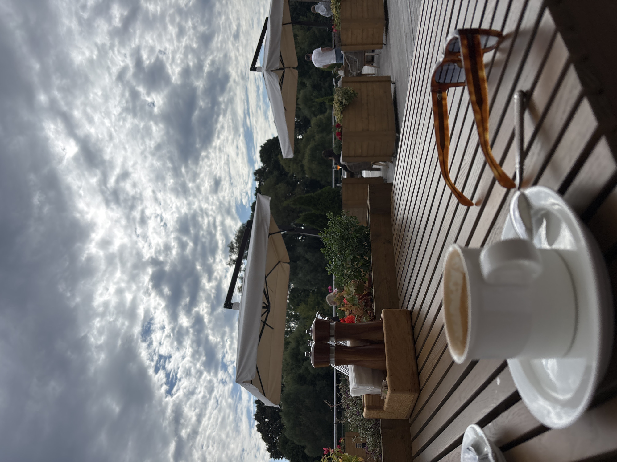 The image depicts an outdoor seating area of a cafe in Zvenigorod, Russia, likely on a cloudy day. In the foreground, a wooden table is set with a white mug of coffee on a saucer, a spoon, and a pair of tortoiseshell sunglasses. To the left, a wooden caddy holds napkins and salt and pepper shakers. In the midground, large wooden planters filled with greenery and red flowers line the edge of the patio. Beyond the planters, umbrellas provide shade over tables where a few patrons are seated. The background features lush green trees and a dramatic, cloudy sky. The overall atmosphere suggests a relaxed outdoor dining experience.
