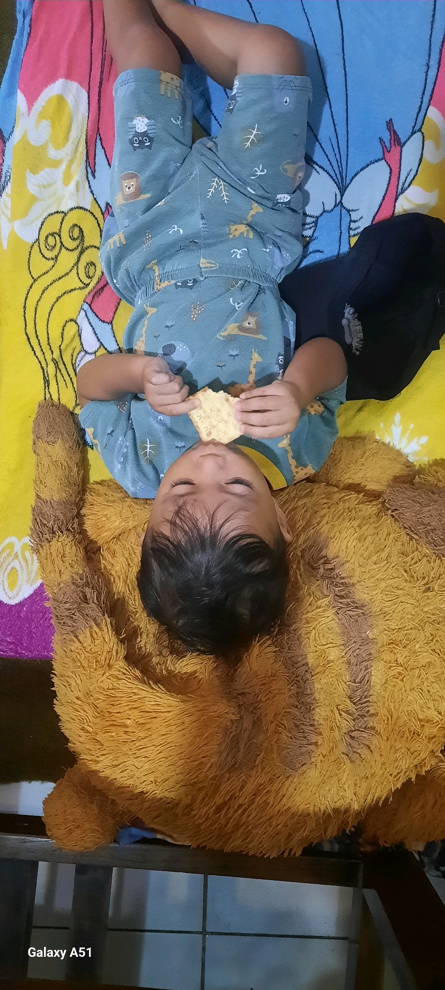In Ploso, Indonesia, a young child lies on their back on a bed. The child is wearing a light blue pajama set adorned with various cartoon animal prints. Their eyes are closed, and they are holding a piece of flatbread in their hands, bringing it towards their mouth. The child is resting on a large, fluffy, brown teddy bear. Behind the child, a colorful bedsheet featuring cartoon characters is visible. The overall scene suggests a moment of rest or snack time for the child.