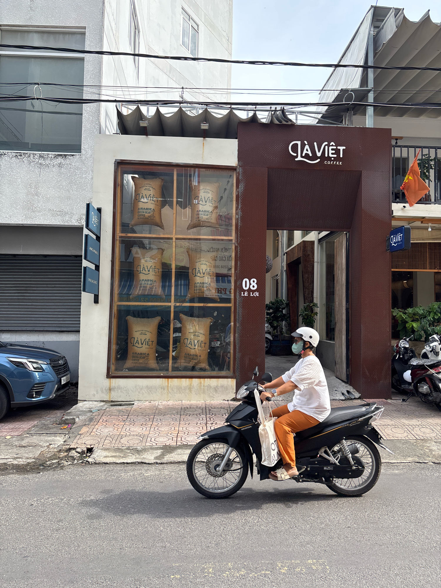 In Nha Trang, Vietnam, a person wearing a white shirt, orange pants, a white helmet, and a light green face mask rides a black Honda motorcycle. The person is carrying a white tote bag on the handlebars. They are riding on a street in front of a building with a brown facade featuring the "CÀ PHÊ VIỆT" logo. Large sacks of coffee beans are visible through the window of the building. To the left of the building, a blue Subaru Forester is parked. The sidewalk is paved with decorative tiles. The overall scene depicts a typical street view in a Vietnamese city.