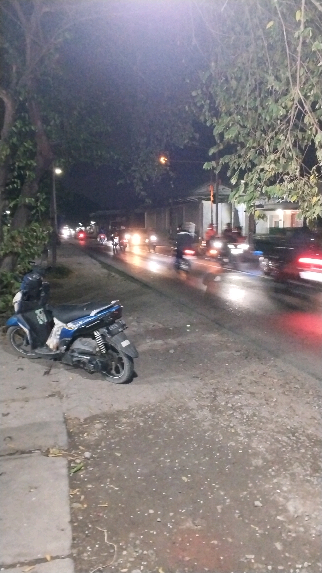 A nighttime street scene in Jetis, Indonesia, depicts a busy road with several motorcycles in motion, indicated by their headlights and taillights. The road is wet, reflecting the lights, suggesting recent rain. On the left side of the road, a blue and black scooter is parked on a dirt and concrete sidewalk. The scooter has a license plate visible. Trees with foliage line the left side of the street, and a street lamp is illuminated. In the background, buildings and more streetlights are visible. The overall atmosphere is that of an active urban environment at night.
