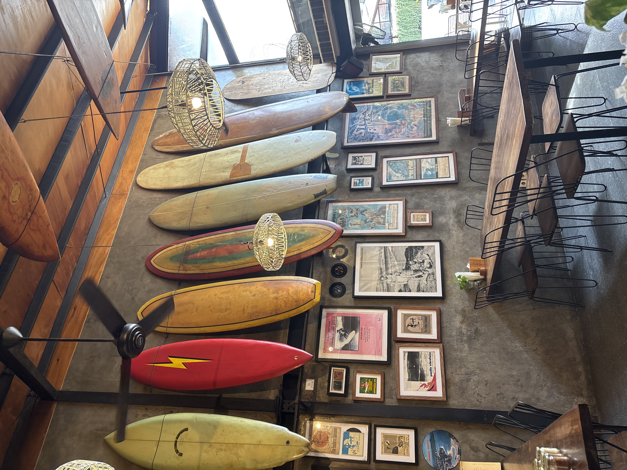 The image shows the interior of a restaurant or café, possibly in North Kuta, Indonesia, judging by the decor. The main elements are the interior walls decorated with a collection of surfboards of various colors and designs, mounted on a grey wall. There are also framed pictures of surfing and surfing-themed items on the wall. Hanging from the ceiling are wicker pendant lights. A ceiling fan is visible amongst the surfboards. The scene includes dining tables and chairs with metal frames, set along the wall, suggesting a dining area. There are no people visible in the shot.