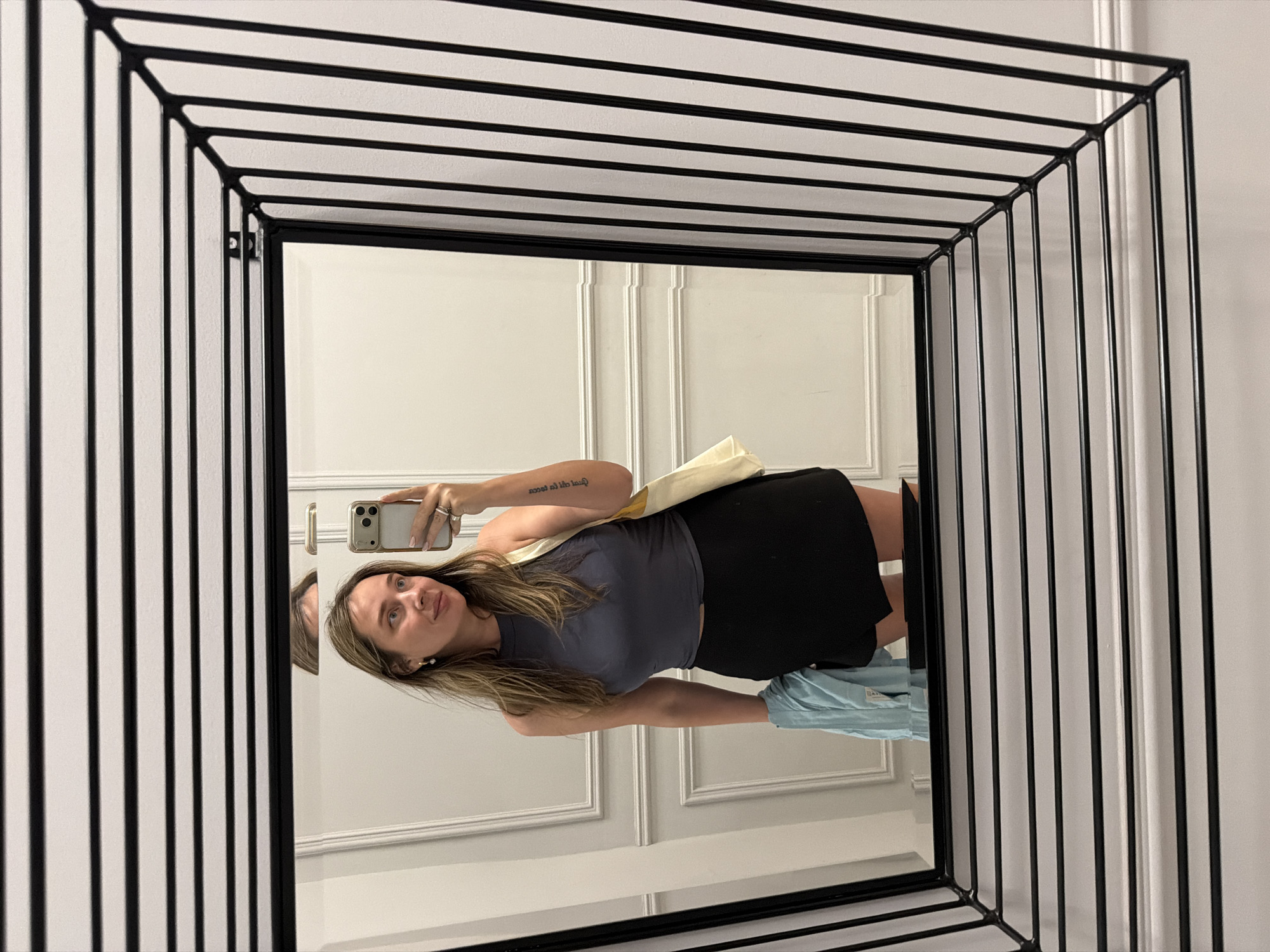 A woman is reflected in a square, black-framed mirror with a decorative cage-like border. She is holding a light-colored smartphone and taking a selfie. She is wearing a dark grey top and a black skirt. A light-colored tote bag hangs from her shoulder, and a light blue garment is partially visible beneath her skirt. The wall behind the mirror is white with a paneled design. The location is likely indoors, but there is no information in the image about it being located in Moscow, Russia.