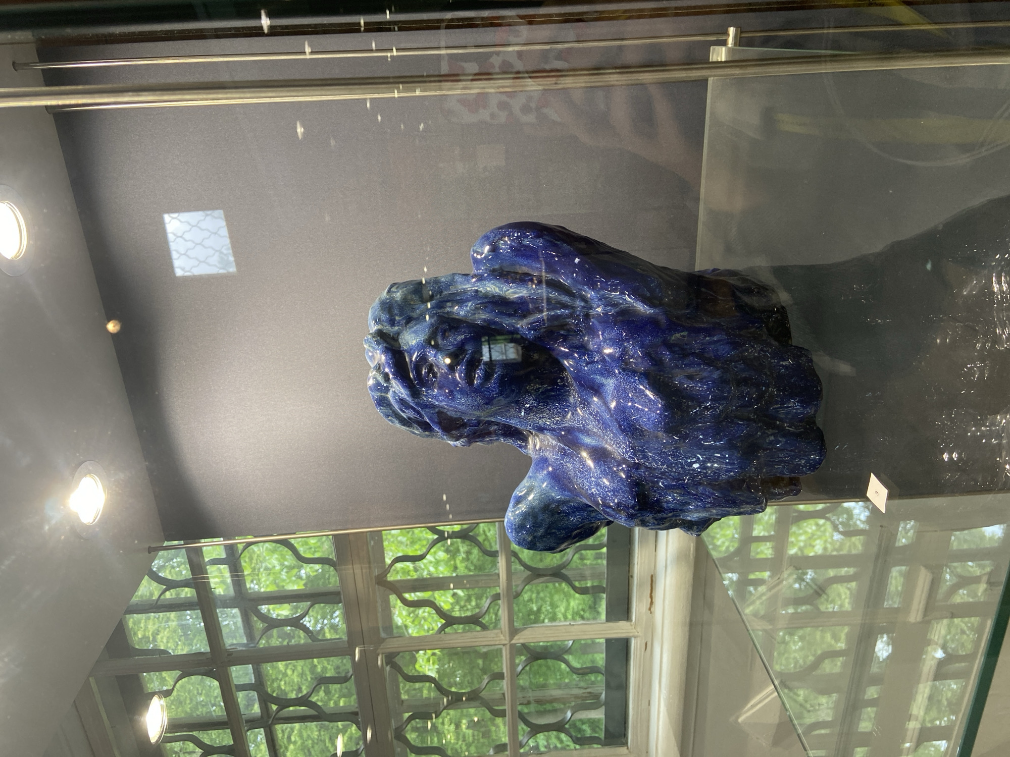 A dark blue, possibly ceramic, sculpture is displayed in a glass case. The sculpture is abstract and its glossy surface reflects light. Behind the sculpture and case is a black wall with a window reflection. The window reflection reveals green foliage outside. Based on the location metadata, this image was taken in Abramtsevo, Russia.