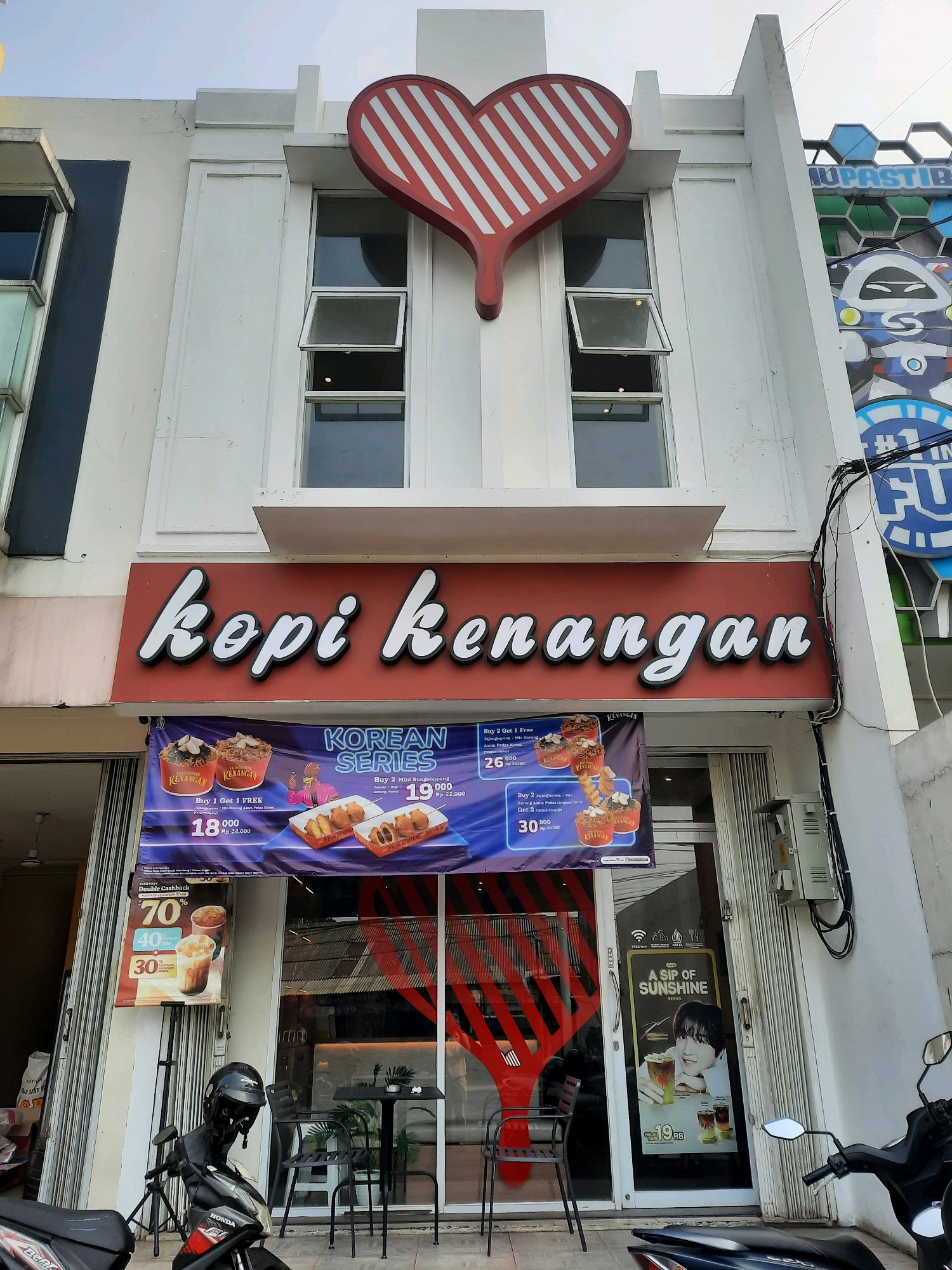 This is an eye-level, full shot of a Kopi Kenangan cafe located on a street in East Jakarta, Indonesia. The building facade is white, with a large red heart-shaped sign with white stripes above the entrance. A prominent red sign with white, cursive lettering reads "kopi kenangan". Below this sign, a large promotional banner displays food and drink offers, including "KOREAN SERIES" and a "DOUBLE CASHBACK" promotion. The storefront features large glass windows and metal folding doors. Inside, tables and chairs are visible, along with potted plants. A motorcycle is parked in front of the cafe. A separate sign for "A SIP OF SUNSHINE SERIES" is placed on the glass door. The sky above is overcast.