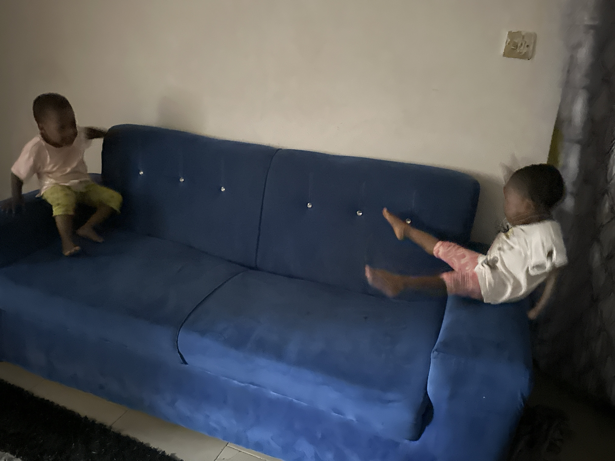 Two young children are on a blue sofa in a room in Narayi, Nigeria. The child on the left is a boy wearing a pink t-shirt and green shorts. He is sitting on the sofa, with his left arm extended towards the backrest. The child on the right is a girl wearing a white t-shirt and pink shorts. She is lying on her stomach on the sofa, with her head turned to the side. The sofa is upholstered in blue fabric and has diamond-shaped buttons on the backrest. The wall behind the sofa is a neutral color, and there is a light switch on the wall. The floor in front of the sofa is covered with a dark rug.