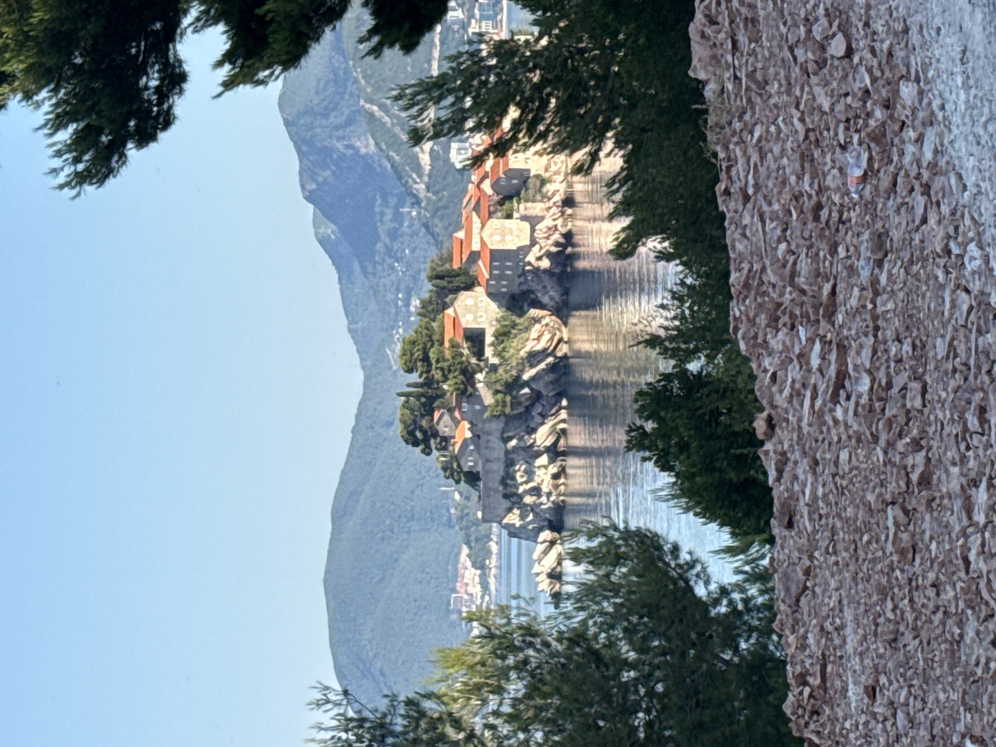 The image captures a scenic view of Sveti Stefan, a small islet and fortified village on the Adriatic coast of Montenegro. The islet is connected to the mainland by a narrow isthmus.
In the foreground, a rocky, uneven terrain is visible, composed of various shades of brown and grey stones. A single plastic bottle lies on the ground in the lower right portion of the image.
Beyond the rocky foreground, lush green pine trees frame the view, with their branches extending into the frame. The trees appear dense and healthy.
The middle ground features the clear, blue waters of the Adriatic Sea, reflecting the sky and the surrounding landscape. The surface of the water shows gentle ripples.
The islet of Sveti Stefan is the focal point of the image. It is a cluster of stone buildings with red-tiled roofs, perched atop a rocky outcrop. The buildings are tightly packed together, creating a picturesque village-like appearance. Some of the buildings have visible windows and architectural details. The islet is surrounded by rocky shores and vegetation.
In the background, rolling green hills rise behind the islet, densely covered with trees. Further in the distance, more layered mountain ranges are visible, fading into a hazy blue. The sky is a clear, pale blue, indicating a sunny day. A few distant buildings and structures can be seen on the coastline to the left of Sveti Stefan.