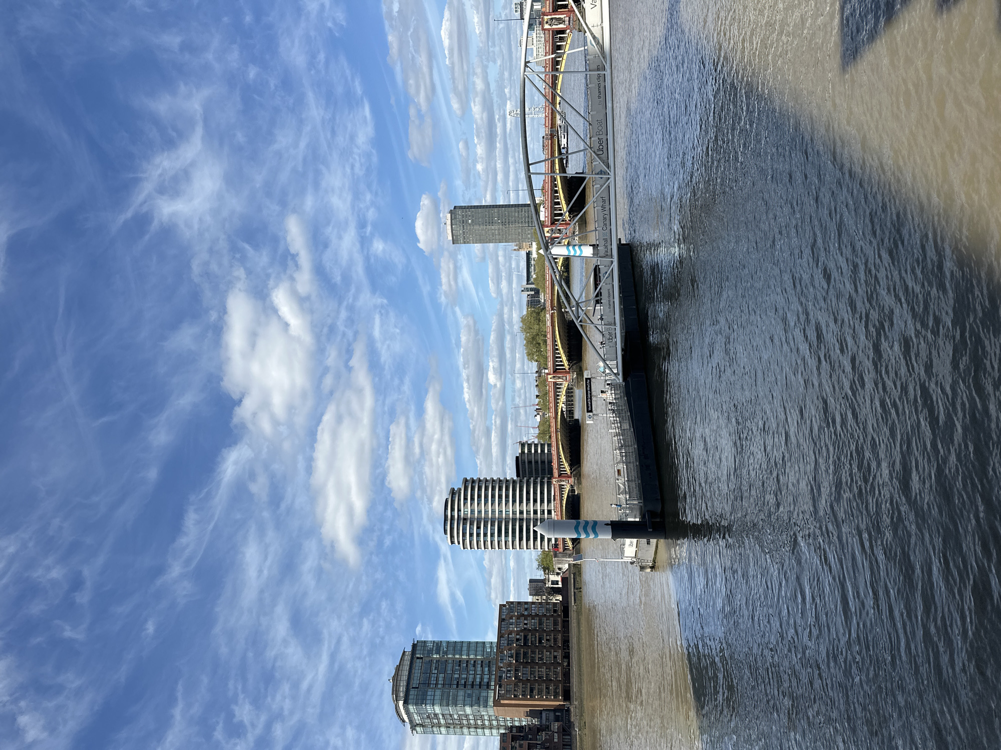 This is a vertical, outdoor shot of the River Thames in London, United Kingdom. The sky is a brilliant blue with scattered white cirrus and cumulus clouds. The river is wide and appears murky with hues of brown and gray. Along the riverbank, there are several modern high-rise buildings. One building stands out with a distinctive spiral design.
A metal structure, likely part of a pier or docking station, is visible on the water. Text "Uber Boat" can be seen written on the pier. A bridge with orange framework is partially visible in the mid-ground.