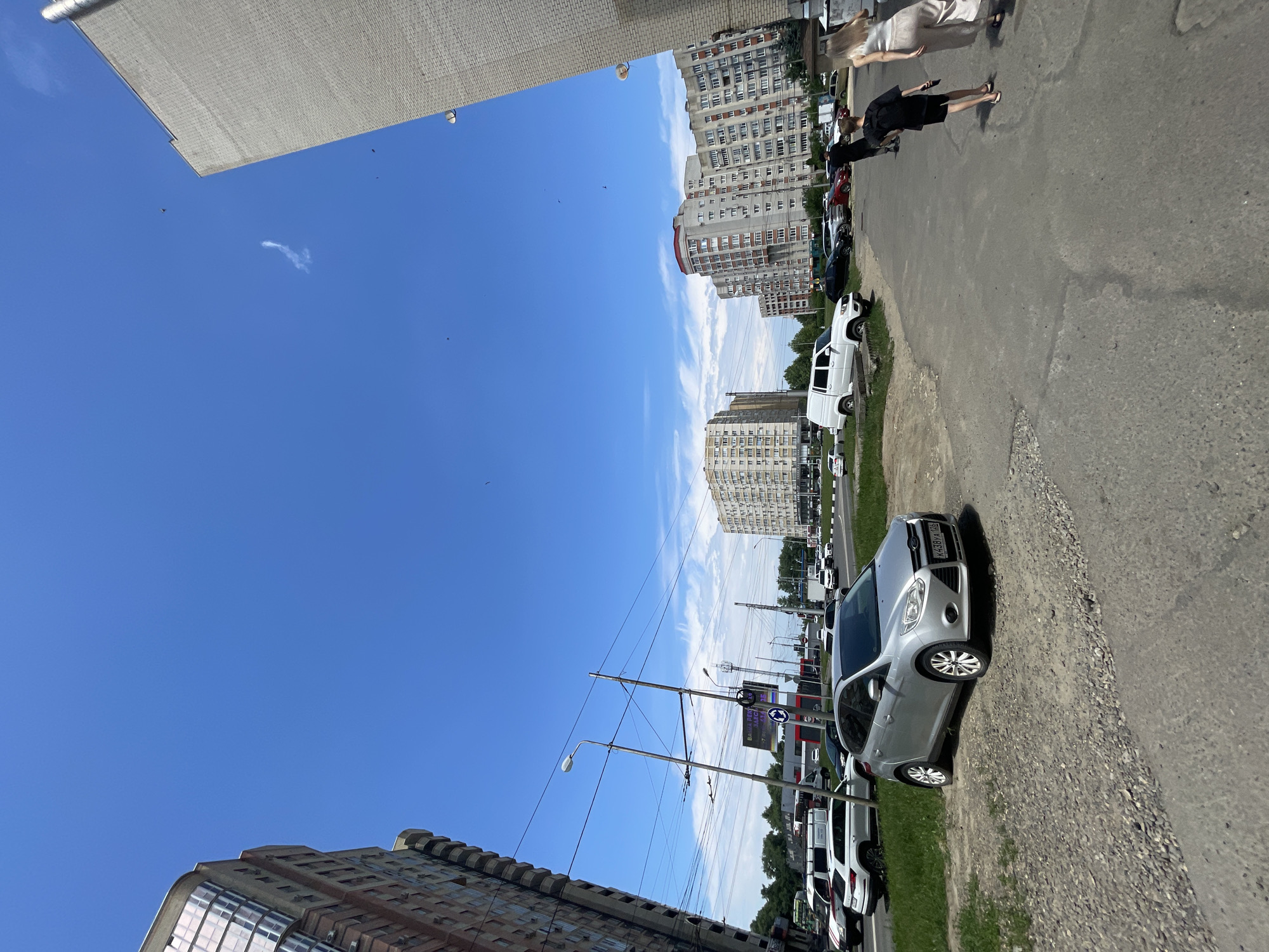 In the city of Stavropol, Russia, on a clear, sunny day, a silver Ford Focus is parked on a gravelly area next to an asphalt road. The car's license plate reads "K438YA 126". To the right of the road, three people are walking away from the camera: a person in a black t-shirt and shorts, a person in a white tank top and light-colored pants, and another person obscured by the two in front. Behind them, a white SUV is parked, and further down the road, several other cars are visible, including white vans and other sedans. Several multi-story apartment buildings are situated in the background, some with red accents on their roofs. Overhead, power lines and streetlights are present, and a billboard with Russian text can be seen. The sky is a bright blue with a few wispy clouds.