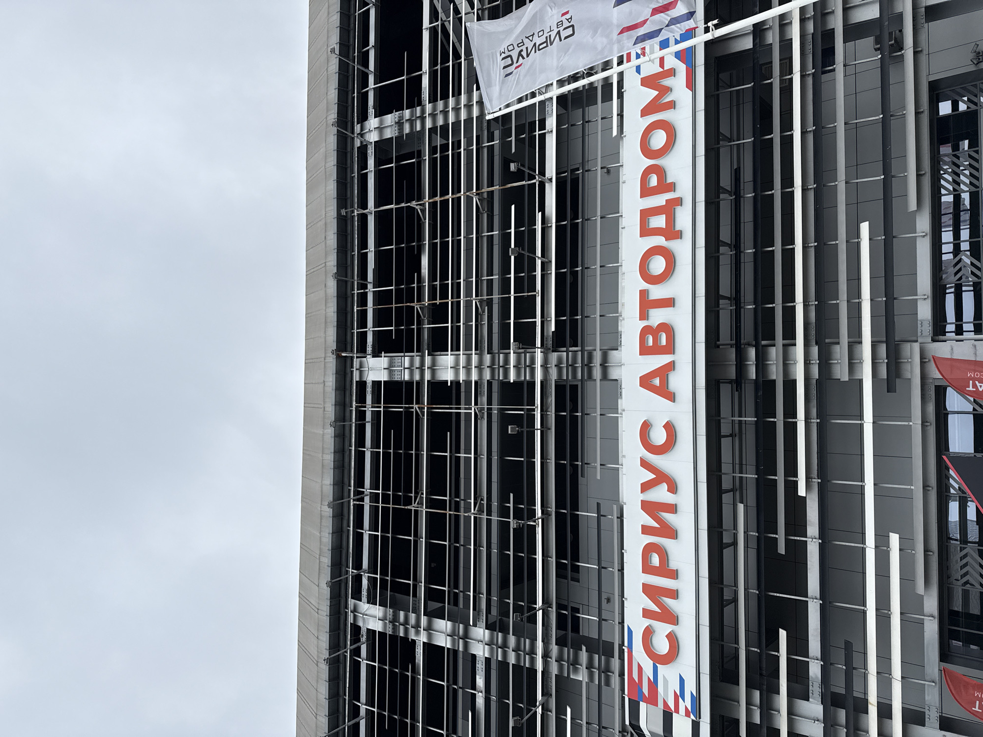 The media file captures a modern building facade from a low, angled perspective, set against an overcast sky. The building features a complex, multi-layered exterior with a prominent grid-like structure of grey metal bars and darker panels or windows behind them. Several vertical white slats also adorn the facade.
A large, vertical sign is affixed to the right side of the building, prominently displaying Cyrillic text in red: "СИРИУС АВТОДРОМ" (SIRIUS AUTODROM). Below "СИРИУС," a stylized graphic in red, white, and blue is visible, resembling a ribbon or wave.
Higher up and to the left of the main sign, a white flag is attached to the building. It also carries the Cyrillic text "СИРИУС АВТОДРОМ," which appears mirrored or reversed, along with a red, white, and blue logo.
The sky is uniformly grey and cloudy, indicating an overcast day. No people or active events are depicted, placing the focus on the building's architecture and branding. The visible text clearly identifies the structure as being associated with "Sirius Autodrom," located in Sirius, Russia.