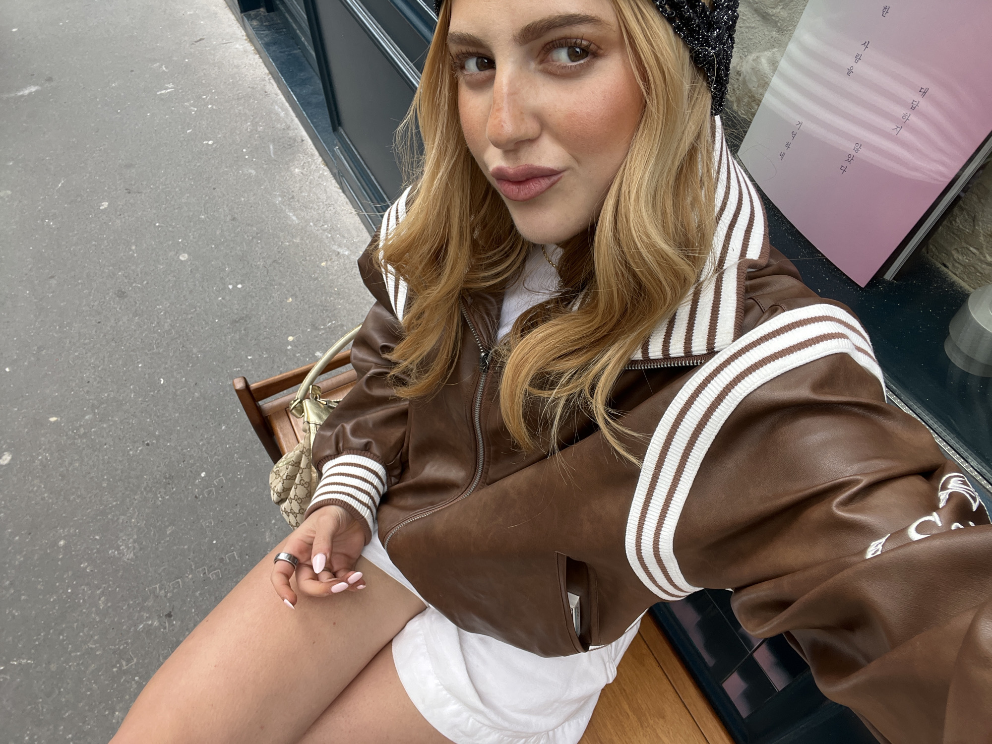 A medium, eye-level selfie shot of a young adult female in an outdoor setting in Paris, France. The female has light skin, long wavy blonde hair, light brown eyes, and medium pink lipstick. She is wearing a brown bomber jacket with white striped sleeves and a white interior shirt. She also wears a glittery dark headband. The female is sitting on a wooden bench, wearing a white skirt or shorts. In her left hand, the female is wearing a silver ring and has light pink painted nails. Behind the female to the left is a patterned designer handbag. The background consists of a grey pavement street, a dark blue window trim, and a book in Korean set against a stone wall. The setting appears to be an outdoor seating area of a shop or café in Paris.
