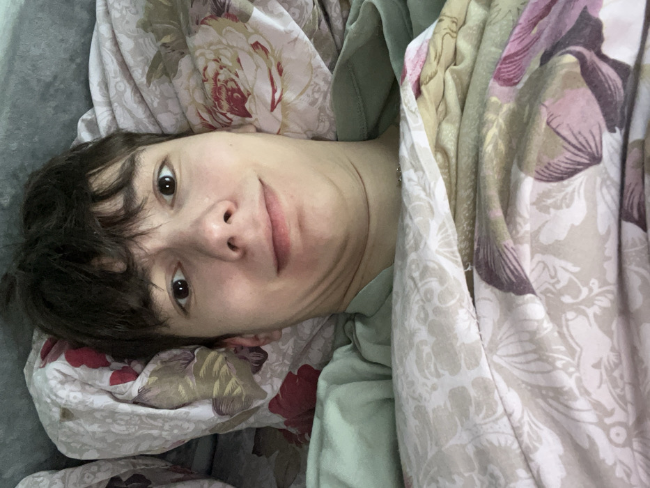 This image depicts a young male individual lying in a bed in Zainsk, Russia. The subject, a young adult with fair skin, short dark brown hair, and dark brown eyes, gazes directly into the camera with a slight, closed-mouth smile. His chin is tucked, creating a visible fold of skin under his jawline. He is wearing a light green t-shirt, visible around his neck and shoulders. The individual is largely covered by patterned bedding, which features large floral designs in shades of cream, beige, pink, red, and purple on a light background. A lighter, possibly grey or white, pillow or headboard is visible behind his head. The scene is illuminated by ambient light, suggesting an indoor bedroom environment.