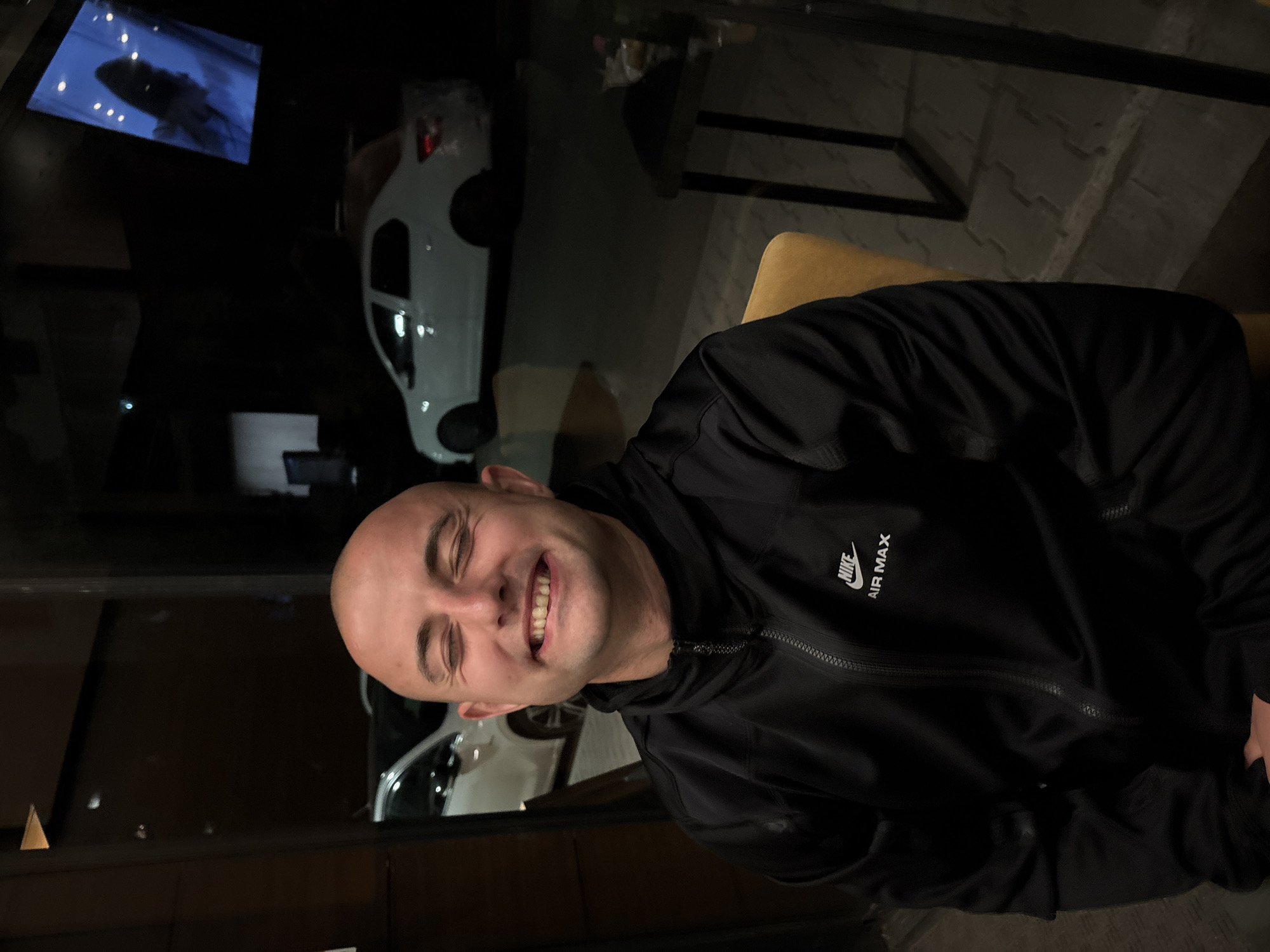 A smiling, bald man wearing a black Nike Air Max jacket is seated. A white car is visible through a window, along with other buildings and a screen displaying an image. The scene appears to be indoors, possibly a restaurant or bar. Based on the image details, the location could be in Sofia, Bulgaria.