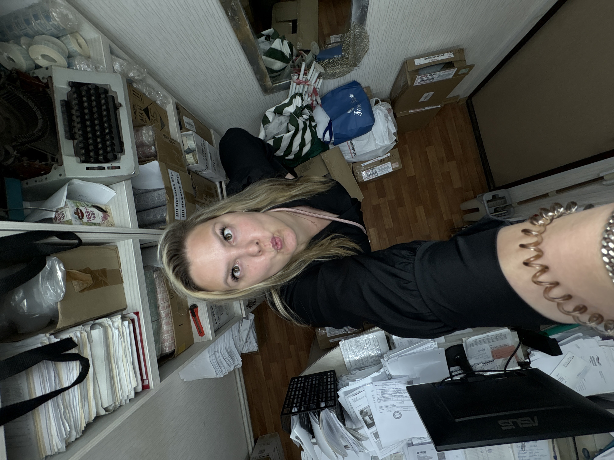 The image is a selfie taken indoors, showing a woman with blonde hair and black clothing. She is looking at the camera with a slightly puckered expression.
Behind the woman is a cluttered environment. A shelf on the left side contains items like rolls of tape, a vintage typewriter, and stacks of files and documents. There are also boxes and a bag.
In the background, there is a mirror reflecting more clutter, including bags and boxes. Further back, the floor appears to be wooden.
To the right of the woman, the scene continues to be cluttered with documents, a black monitor with the brand name "ASUS" visible, and other miscellaneous items on a desk. Text and brands like "PECO" and "BUS" are visible on some of the documents and boxes.
The OCR of the image includes "иван чай" (Ivan tea), "ИП Мишихин 60 р." (IP Mishikhin 60 rubles), "ИП Мащихии 50 р-" (IP Maschikhin 50 rubles), "Dokumenty 45" (Documents 45), and "Noviy" (Новый).